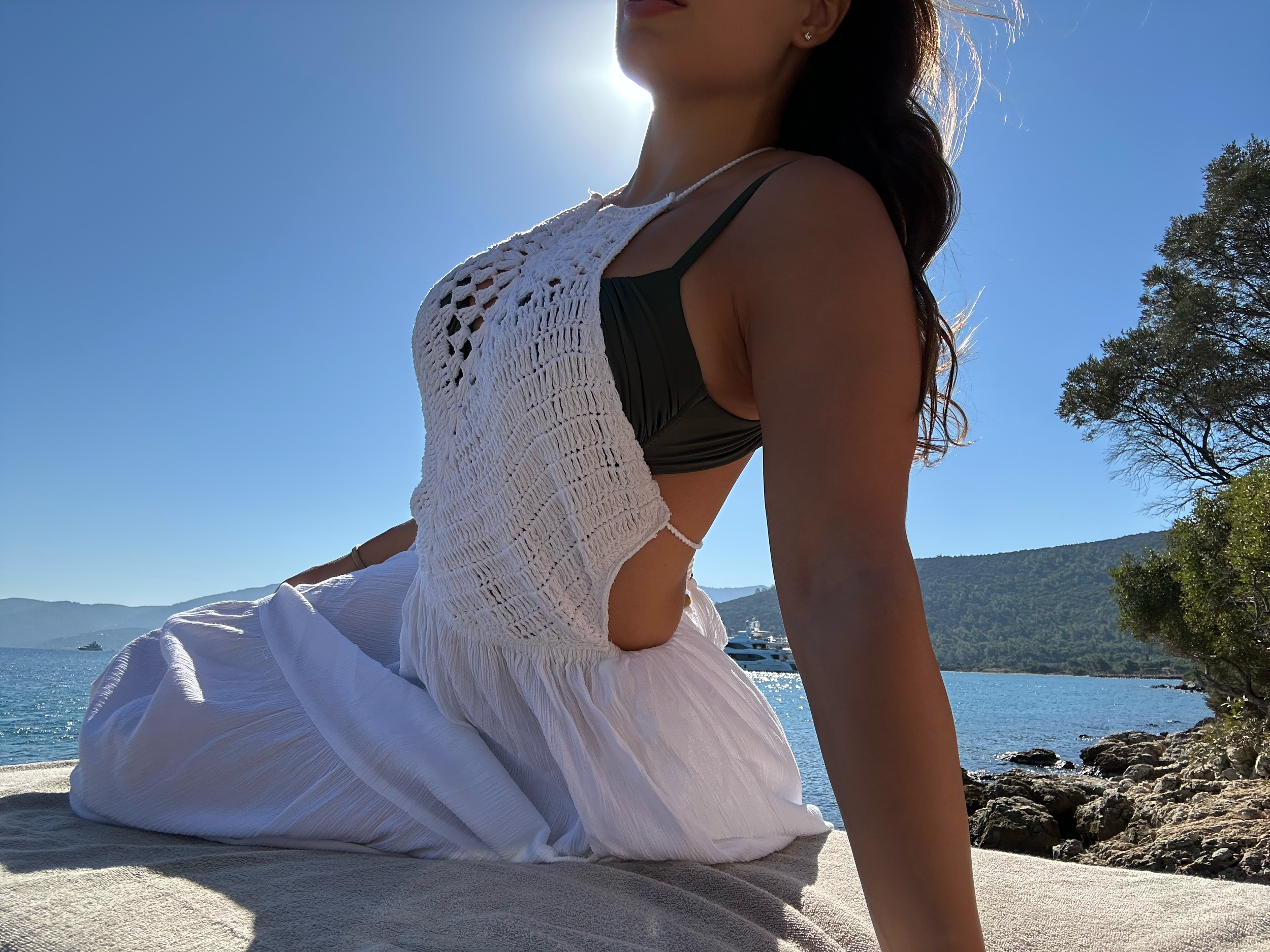 Arabella Blair, Elite Travel Companion Escort Model, sitting outdoors at Amanruya, an Aman resort, next to the sea, with boats, trees, and mountains in the background, backlit by the sun.