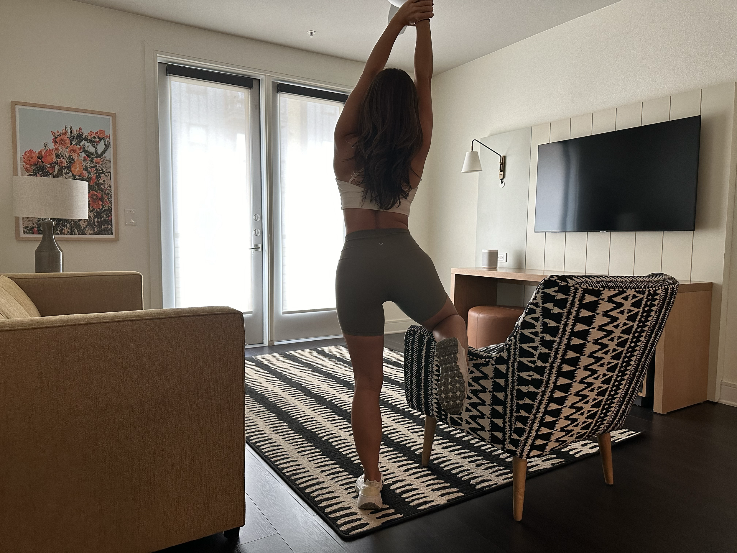 Arabella Blair, Independent Escort in London, stretching in a living room with a striped rug, patterned armchair, television, and sliding glass doors.