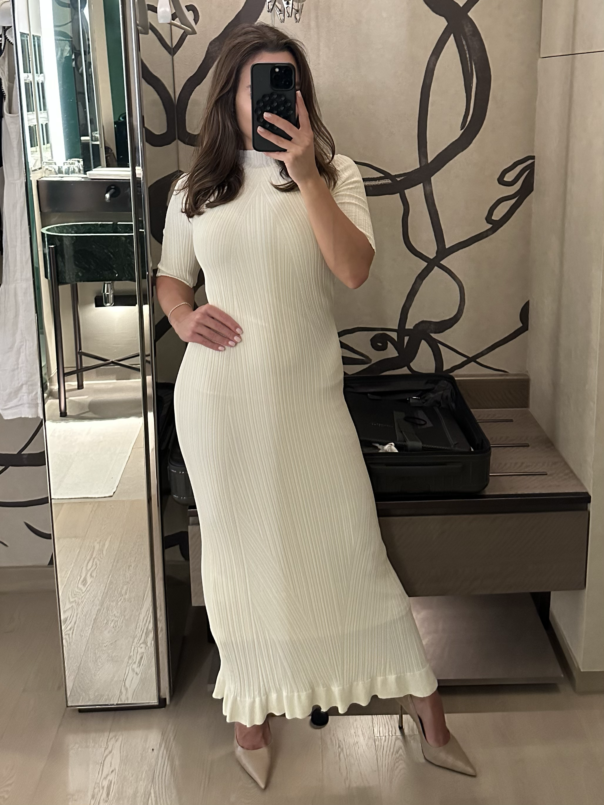 Arabella Blair, London’s GFE Escort, in a cream-colored, ribbed, midi dress with ruffled hem, wearing beige high heels, taking a mirror selfie in a modern room with artwork on the walls.