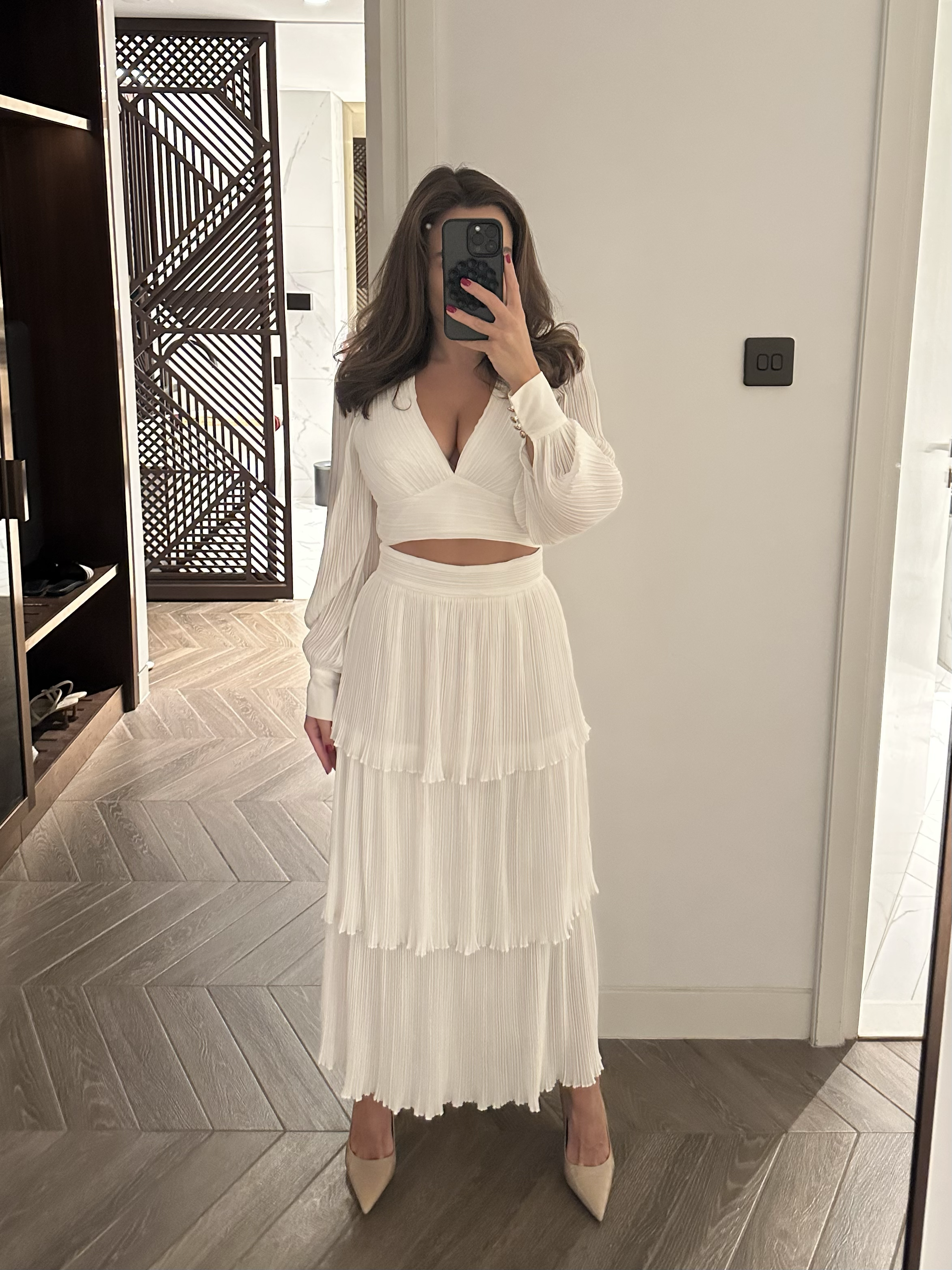 Arabella Blair, the best GFE Escort in London, wearing a white pleated, tiered skirt and matching long-sleeved top, taking a mirror selfie in a room with wooden flooring and a decorative wooden partition.