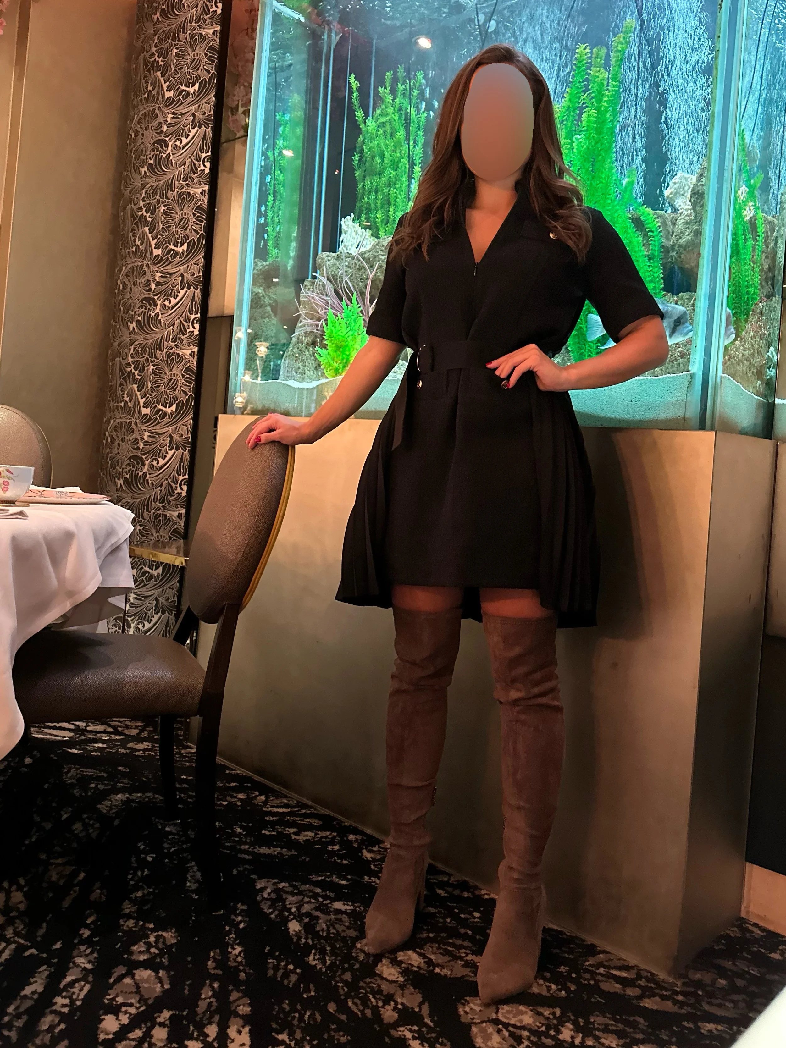 Arabella Blair, London’s most requested Dinner Date Escort, standing in front of an aquarium with green aquatic plants and rocks. She is wearing a black dress and over-the-knee brown boots.