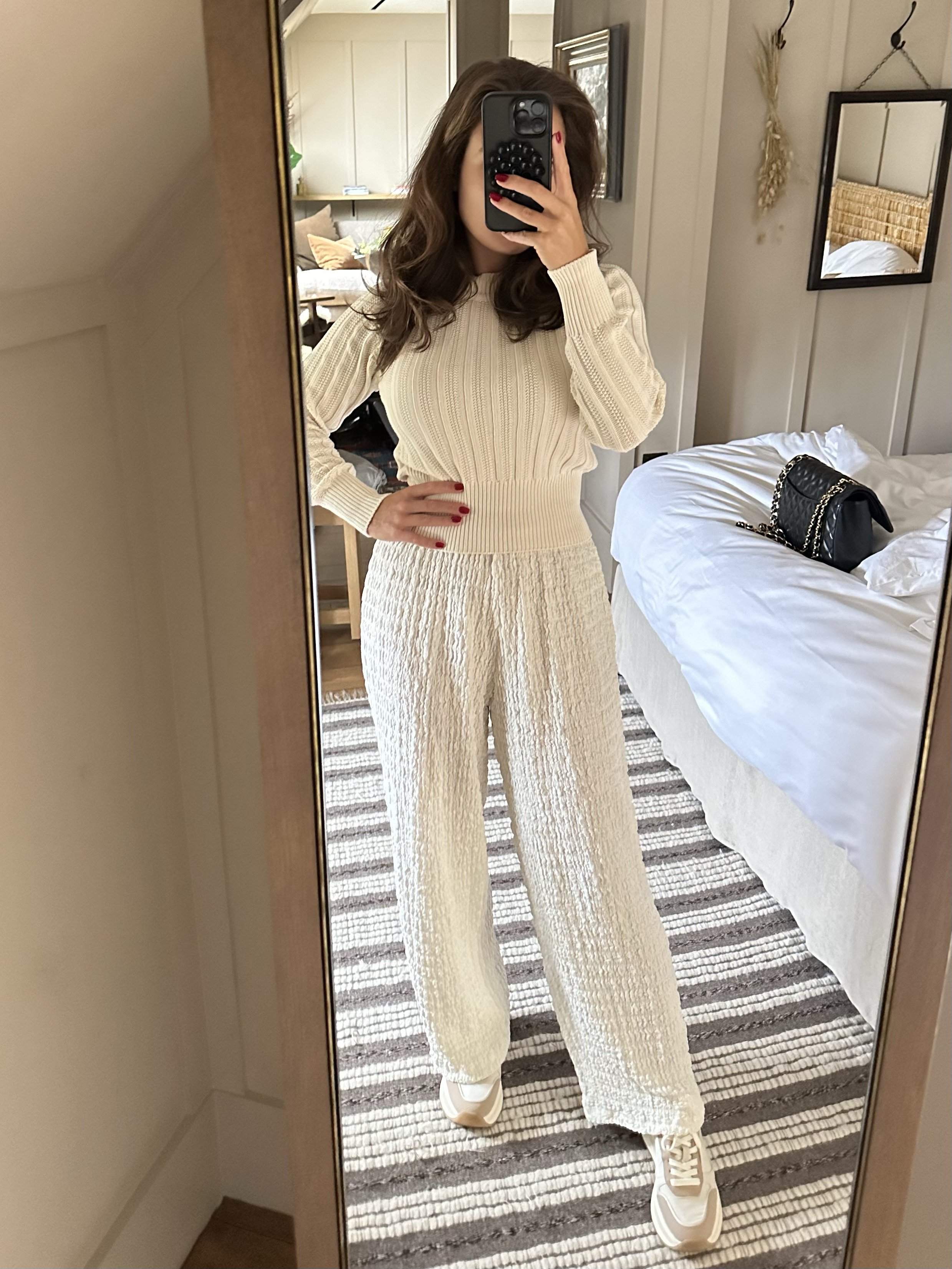 Arabella Blair, Heckfield Place’s most requested Girlfriend Experience Escort, taking a mirror selfie dressed in a cream-colored, ribbed knit sweater and matching loose-knit pants, wearing white sneakers, with a black purse on the bed behind her.
