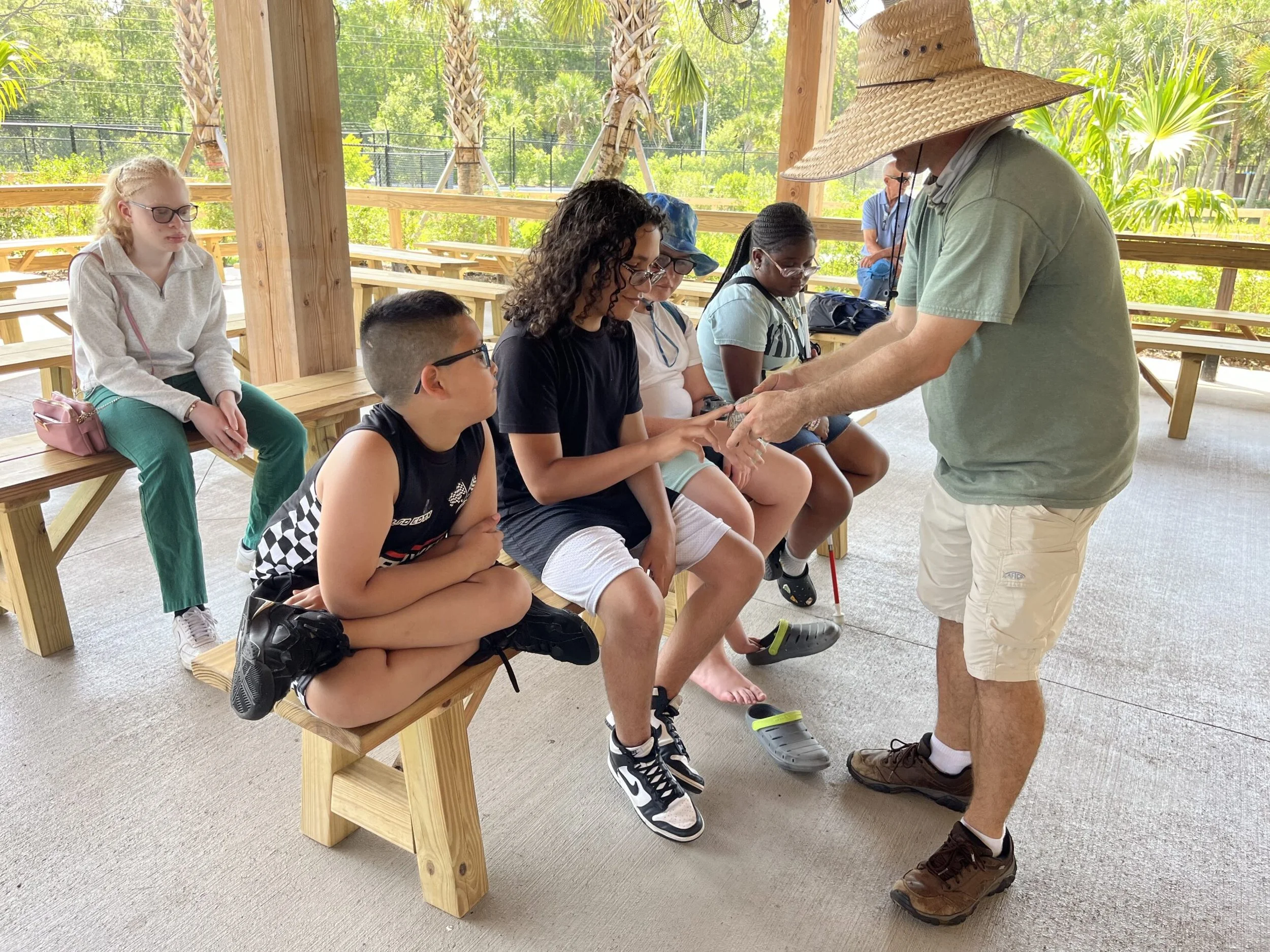 Young clients of Lighthouse for the Blind of the Palm Beaches interact with an animal ambassador at Busch Wildlife Sanctuary at an earlier event. They are seated on benches under a pavilion interacting with an educator and small tortoise.