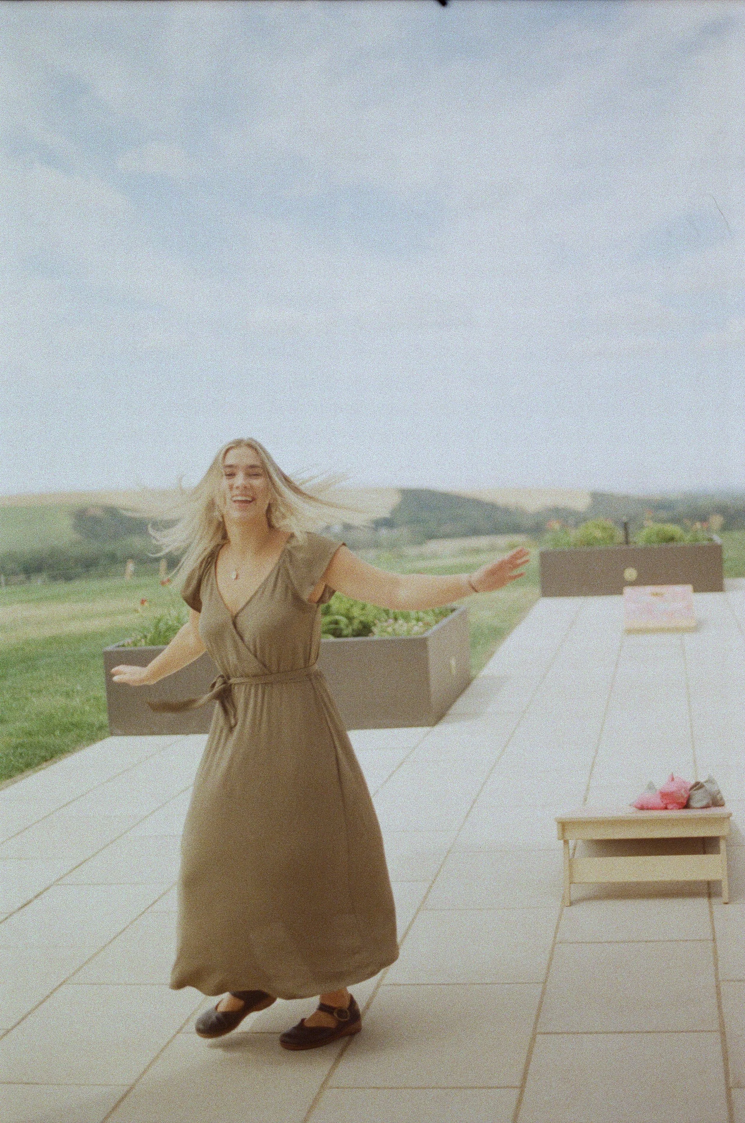 A woman dancing outdoors on a patio with green fields and hills in the background, smiling and wearing a brown dress.