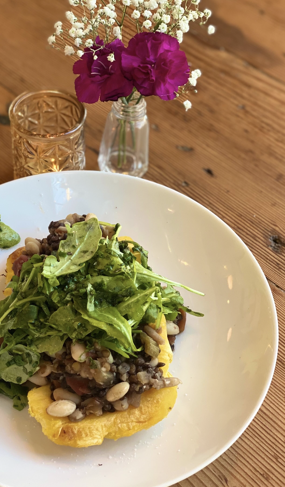 A white plate with a baked dish topped with a fresh green salad on a wooden table, a small vase with purple and white flowers, and a textured amber glass of water in the background.