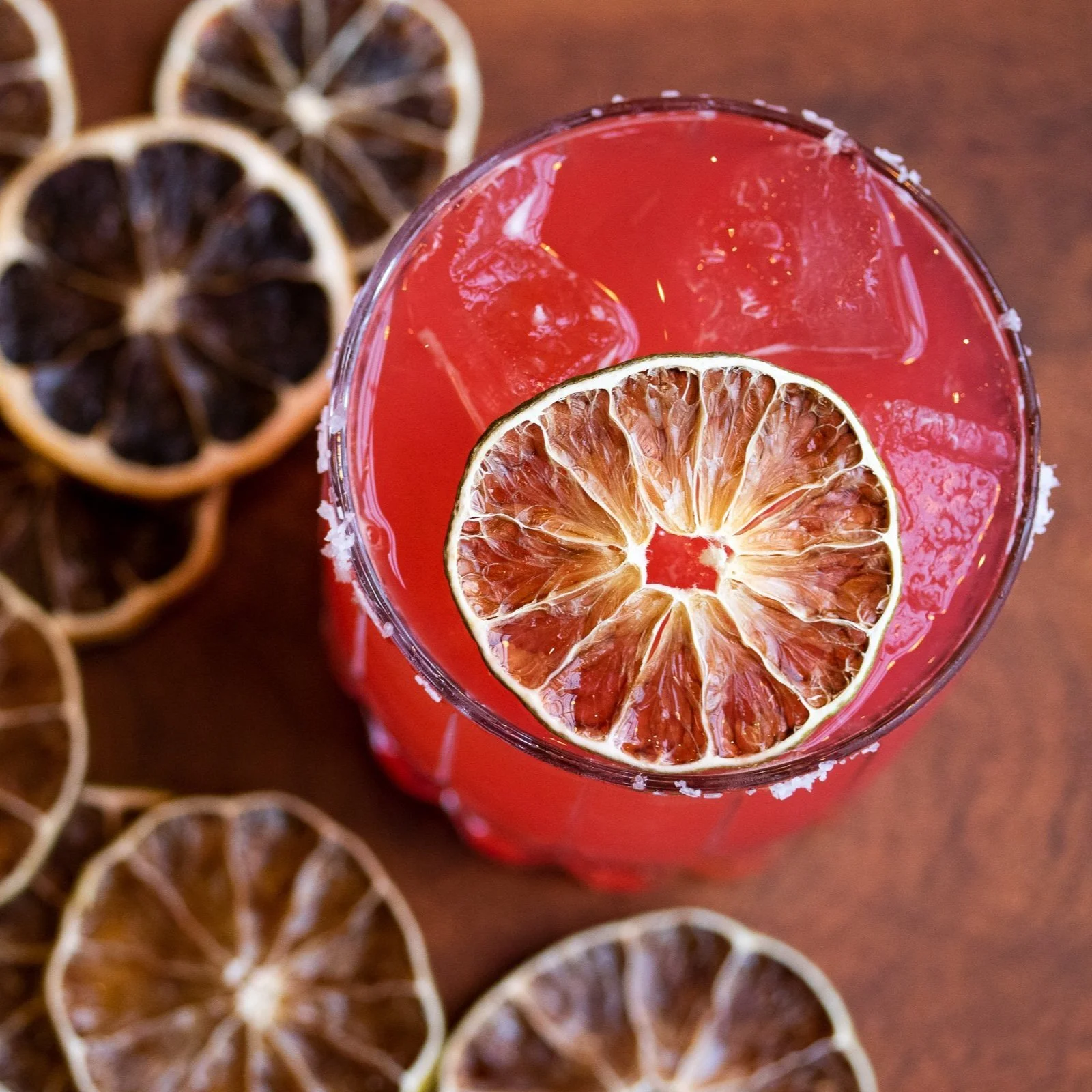 A glass of red beverage with a dried lemon slice on top, surrounded by dried lemon slices on a wooden surface.