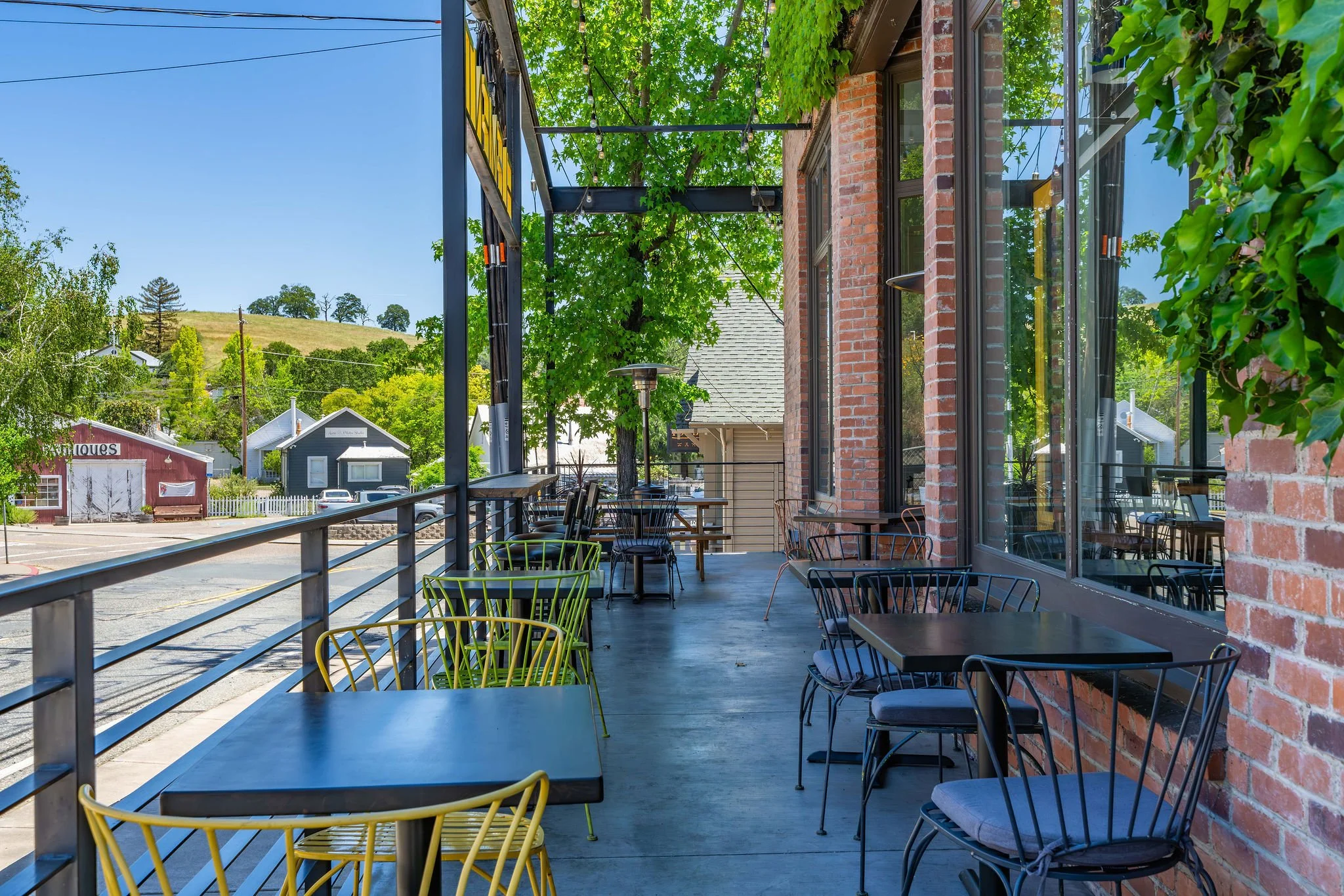 Outdoor seating on a restaurant or café patio with black tables, colorful chairs, brick wall, large windows, and greenery, overlooking a street and hillside beyond.