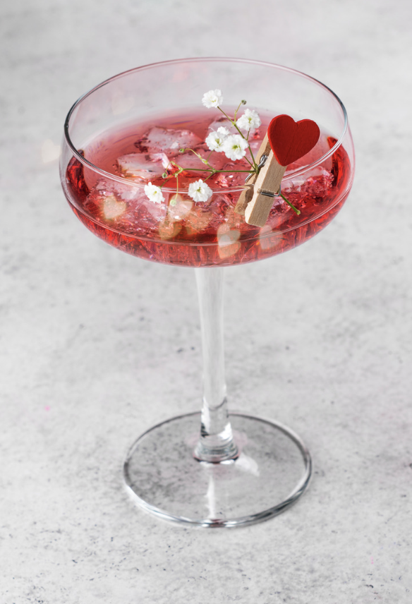 A martini glass with a pink beverage, garnished with white flowers, a small red heart, and a clothespin, sitting on a white surface.