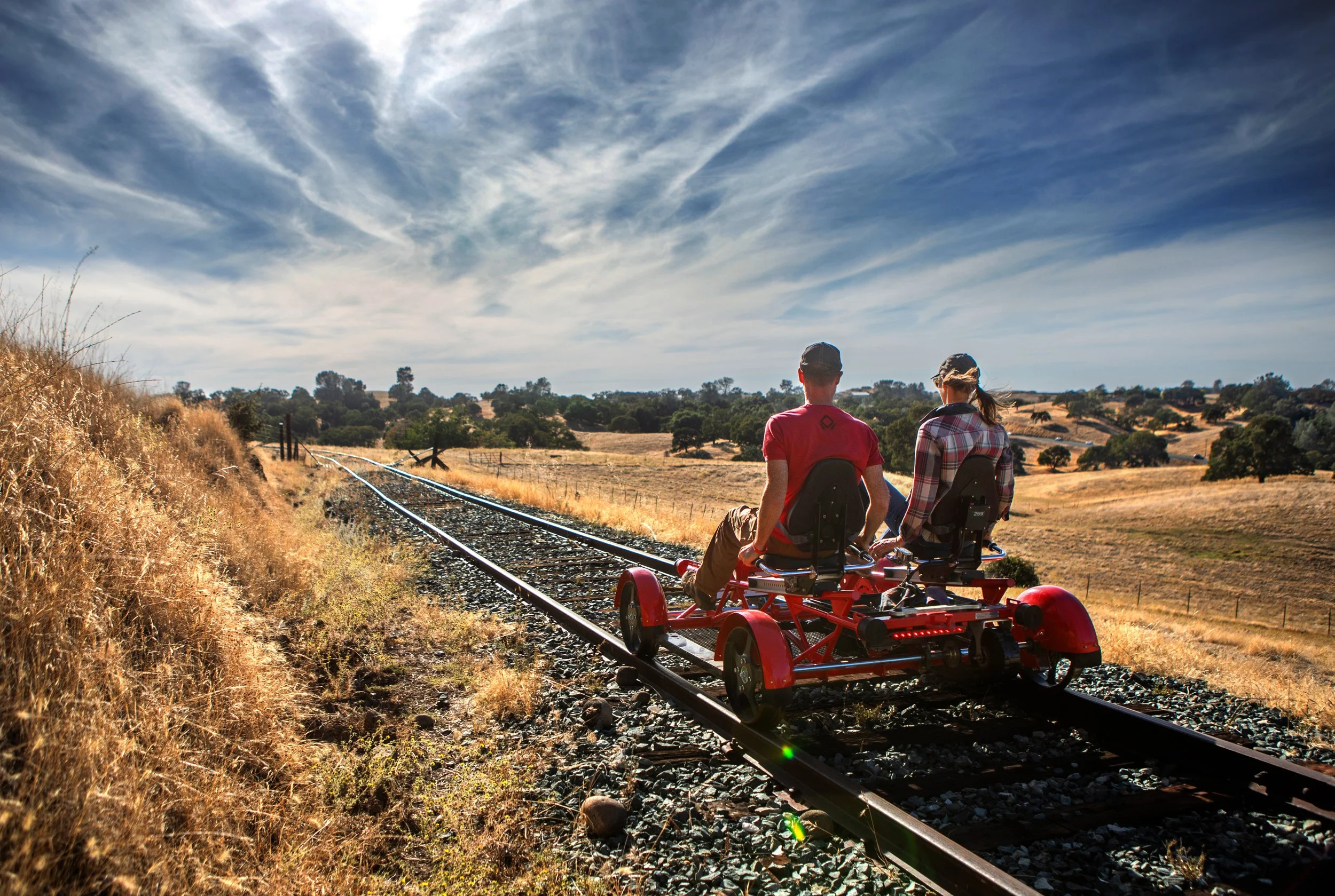 Two people riding a red rail bike along a rural train track through a dry, grassy landscape under a partly cloudy sky.