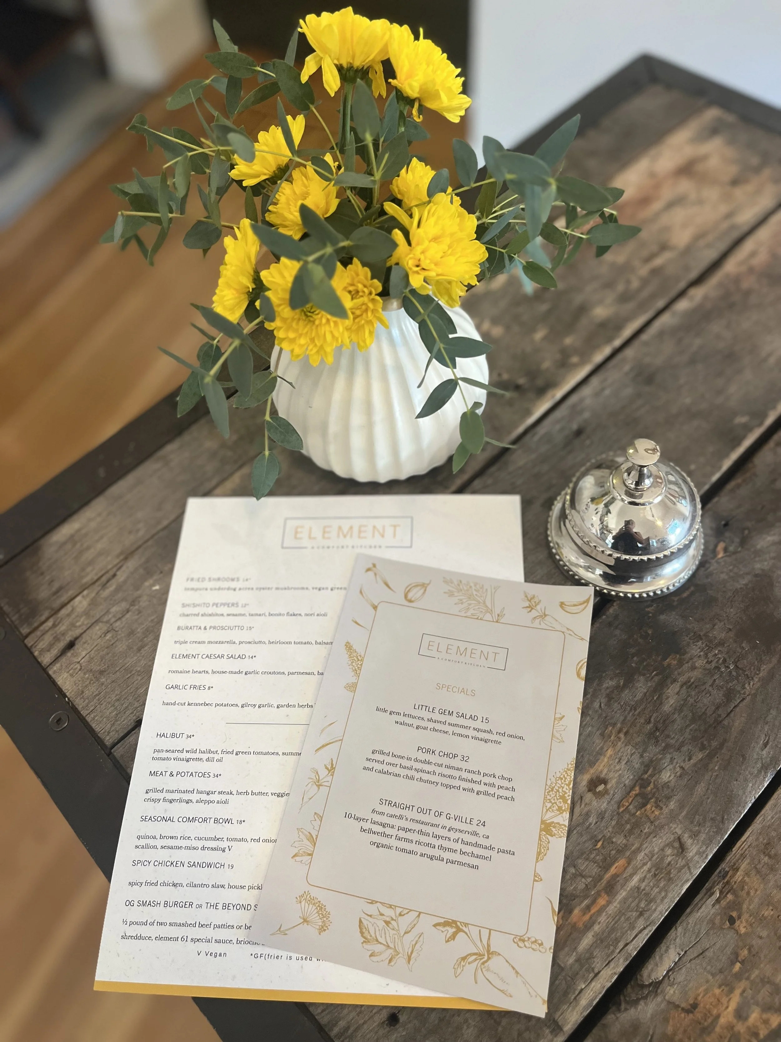 A bouquet of yellow flowers and green leaves in a white shell-shaped vase on a rustic wooden table, with two menus and a small silver container nearby.