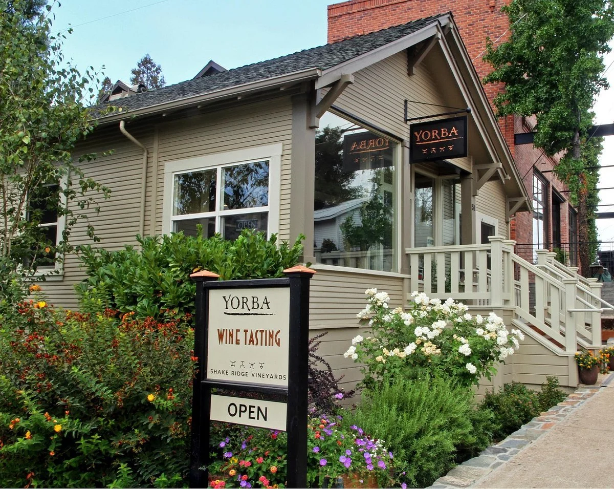 Exterior of a beige building with a staircase and a sign that reads 'YORBA WINE TASTING' with a smaller sign indicating the business is open, surrounded by lush greenery and flowers.