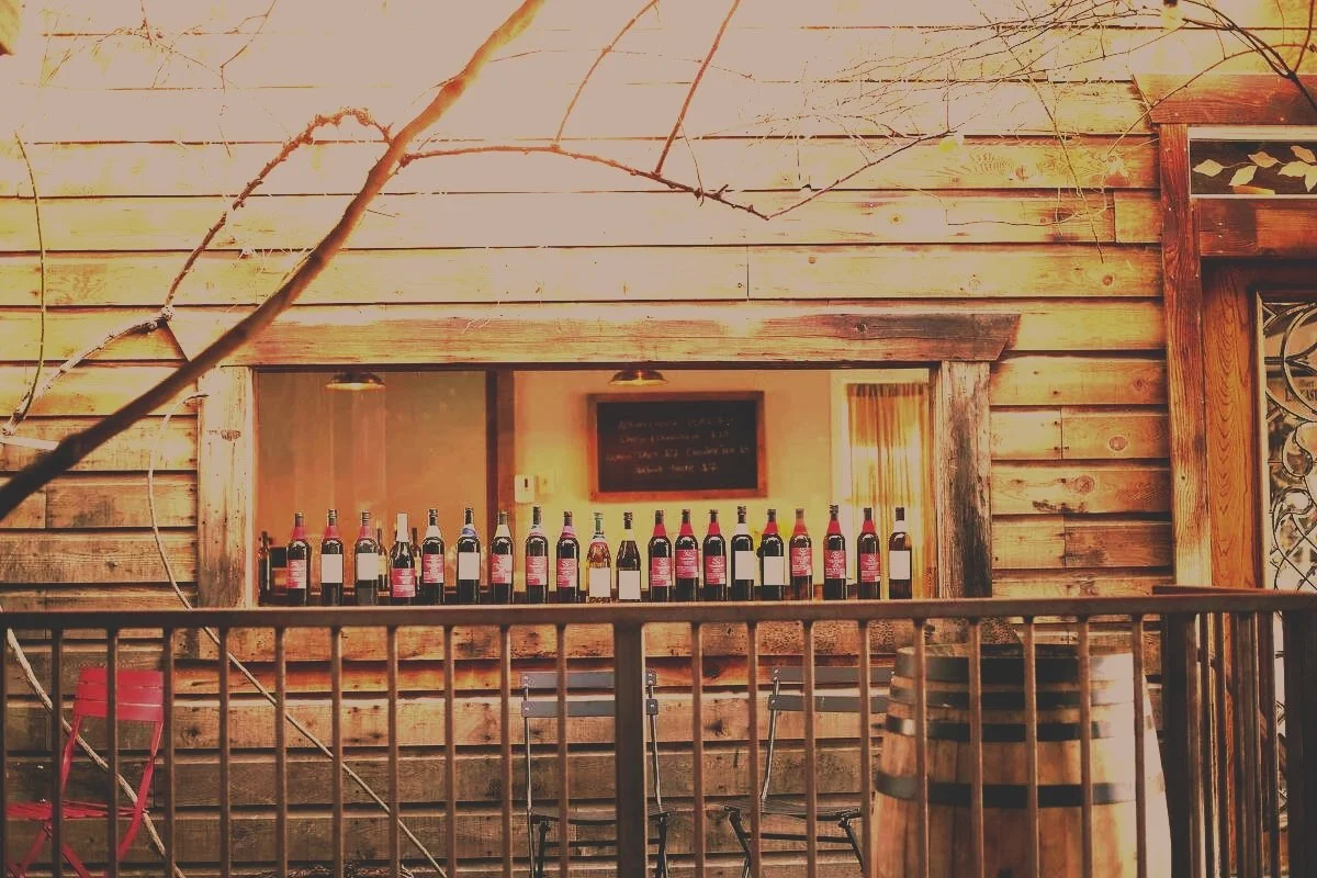 A row of wine bottles displayed on a wooden counter or shelf in a rustic setting with wooden walls, a chalkboard, and a wooden wine barrel in the foreground.
