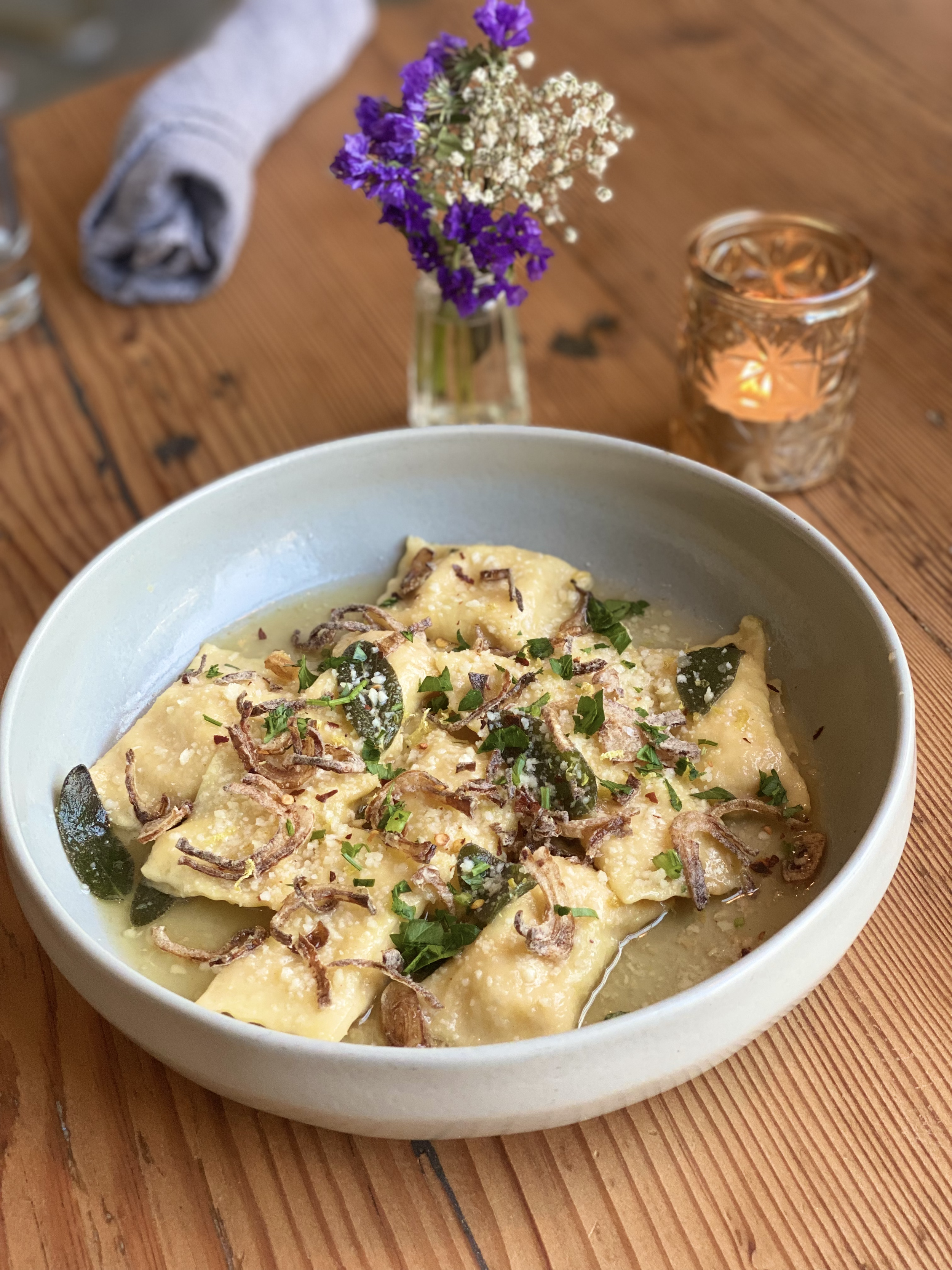 A bowl of ravioli pasta topped with chopped herbs, fried shallots, and freshly grated cheese, served with a small flower vase and a candle holder on a wooden table.