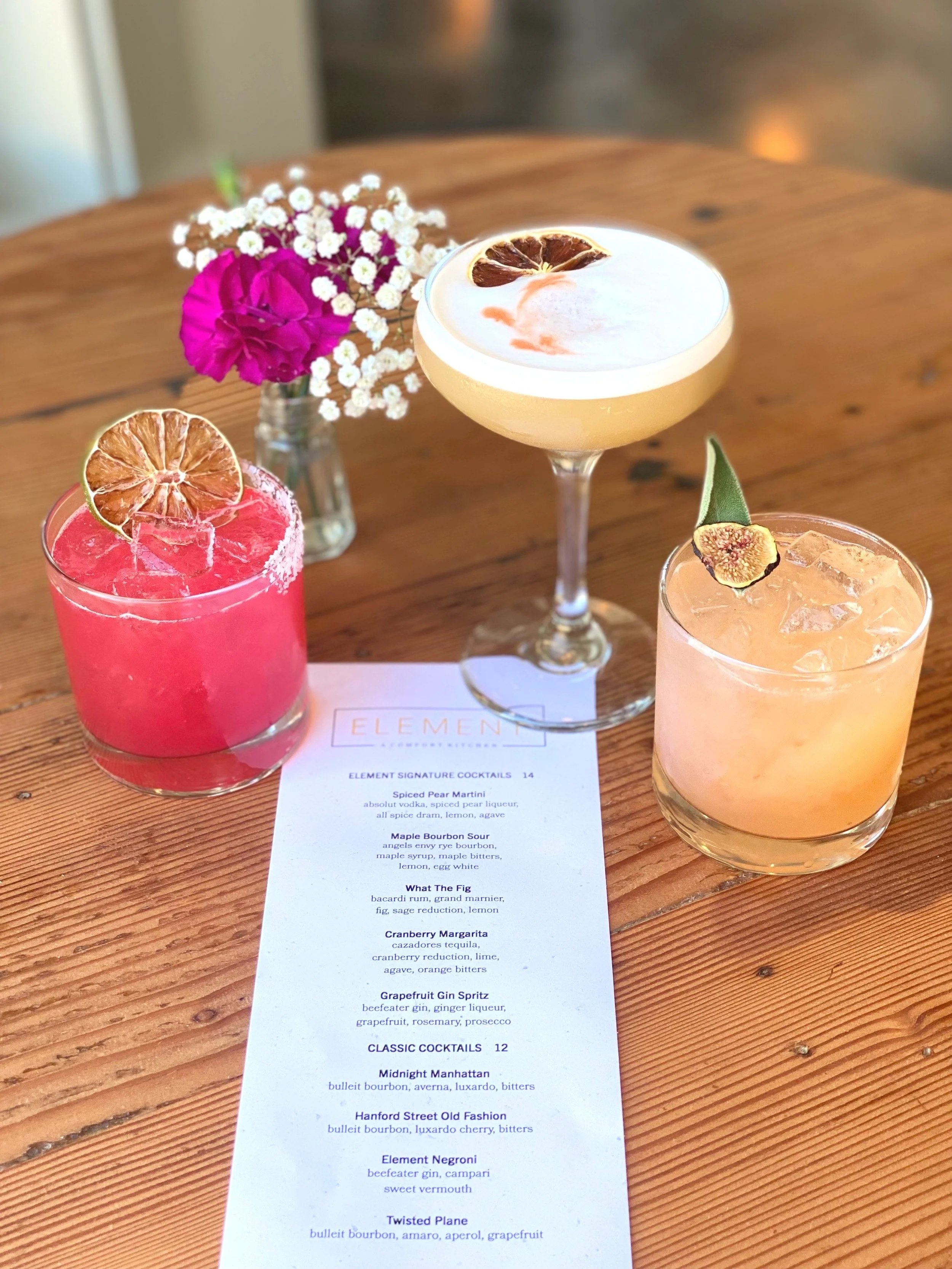 Three cocktails on a wooden table with a flower arrangement in the background. One yellow cocktail in a coupe glass garnished with a dried lemon slice, one pink cocktail in a rocks glass with a dried lemon slice, and one orange cocktail in a rocks gl