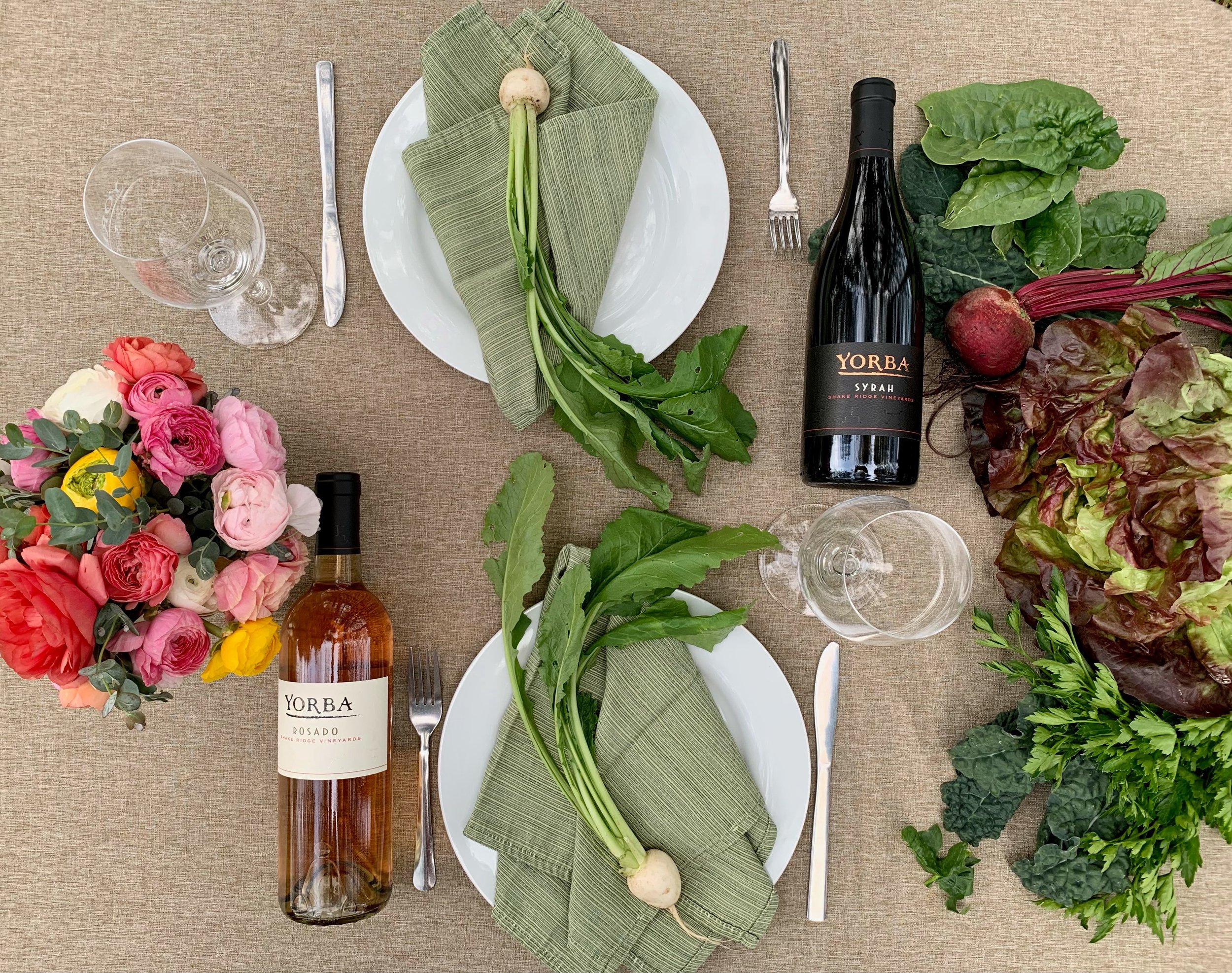 A table set for two with plates, silverware, green napkins, and wine glasses. Two bottles of wine and a bouquet of pink, red, yellow, and white flowers are present, along with fresh vegetables including beets, spinach, and lettuce.
