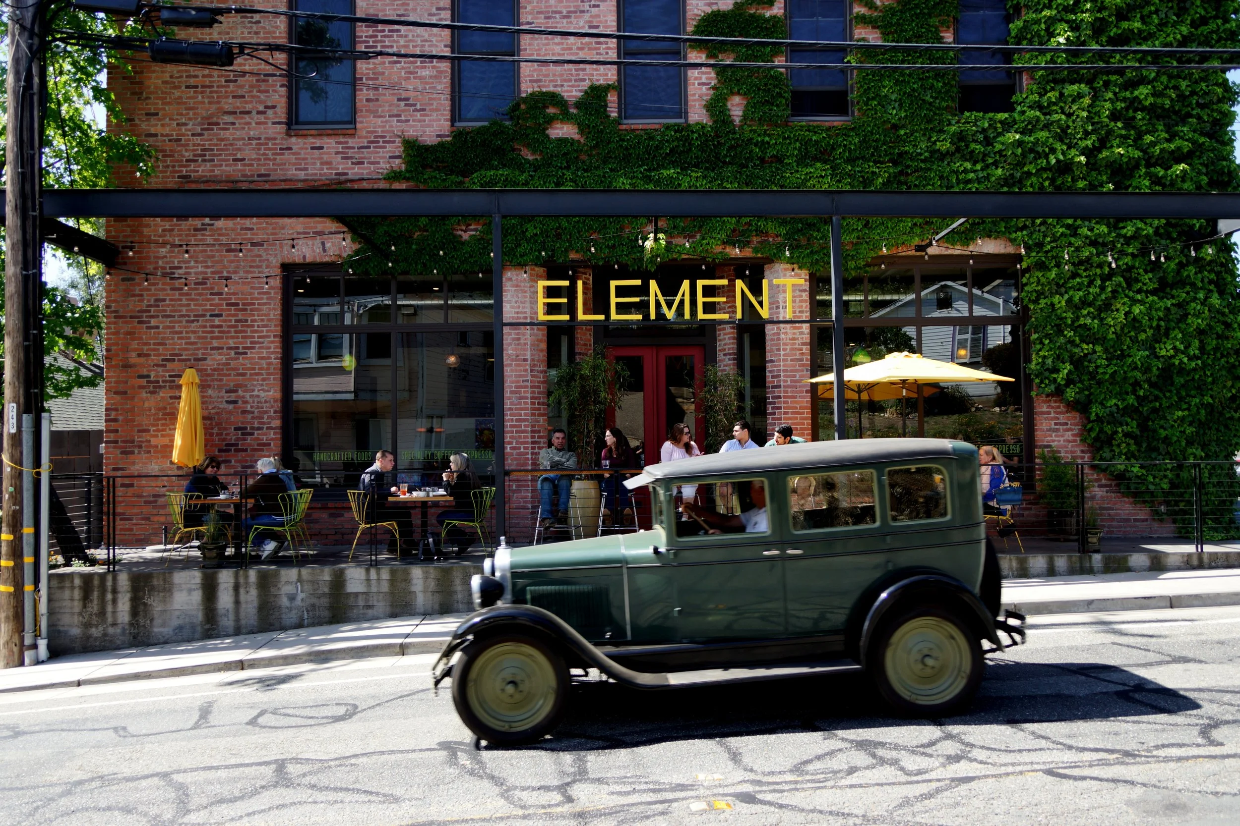 A vintage green car driving past a brick building with a sign that reads 'ELEMENT' and outdoor seating with people dining under yellow umbrellas.