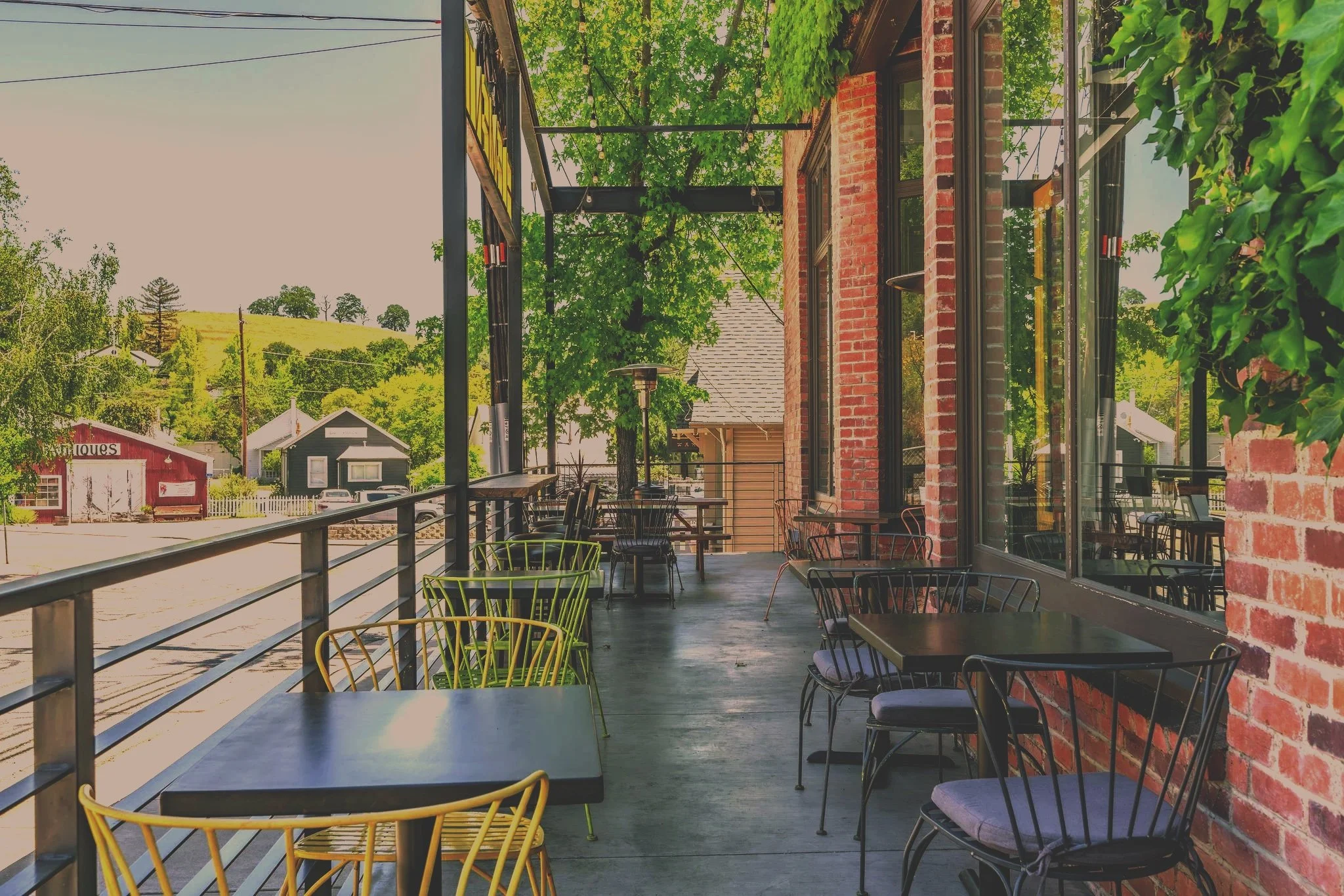 Empty outdoor patio with black tables and chairs, brick building, green leafy trees, and hillside in background