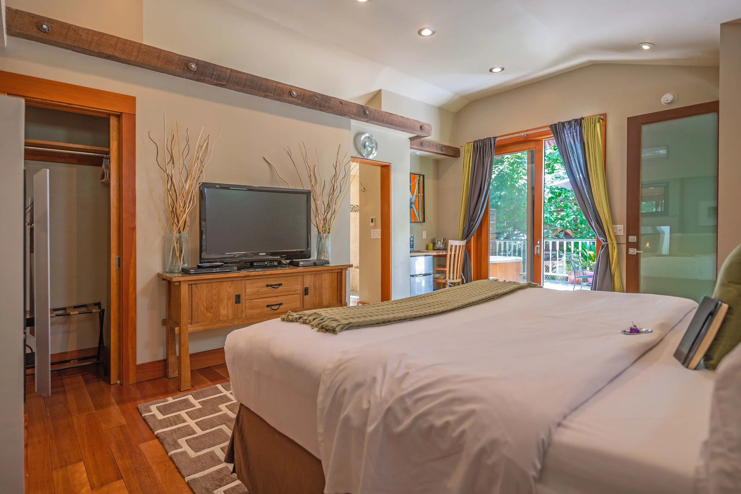 A cozy bedroom with wooden flooring, a large bed with white linens, a flat-screen TV on a wooden dresser, and a balcony door with curtains leading outside.