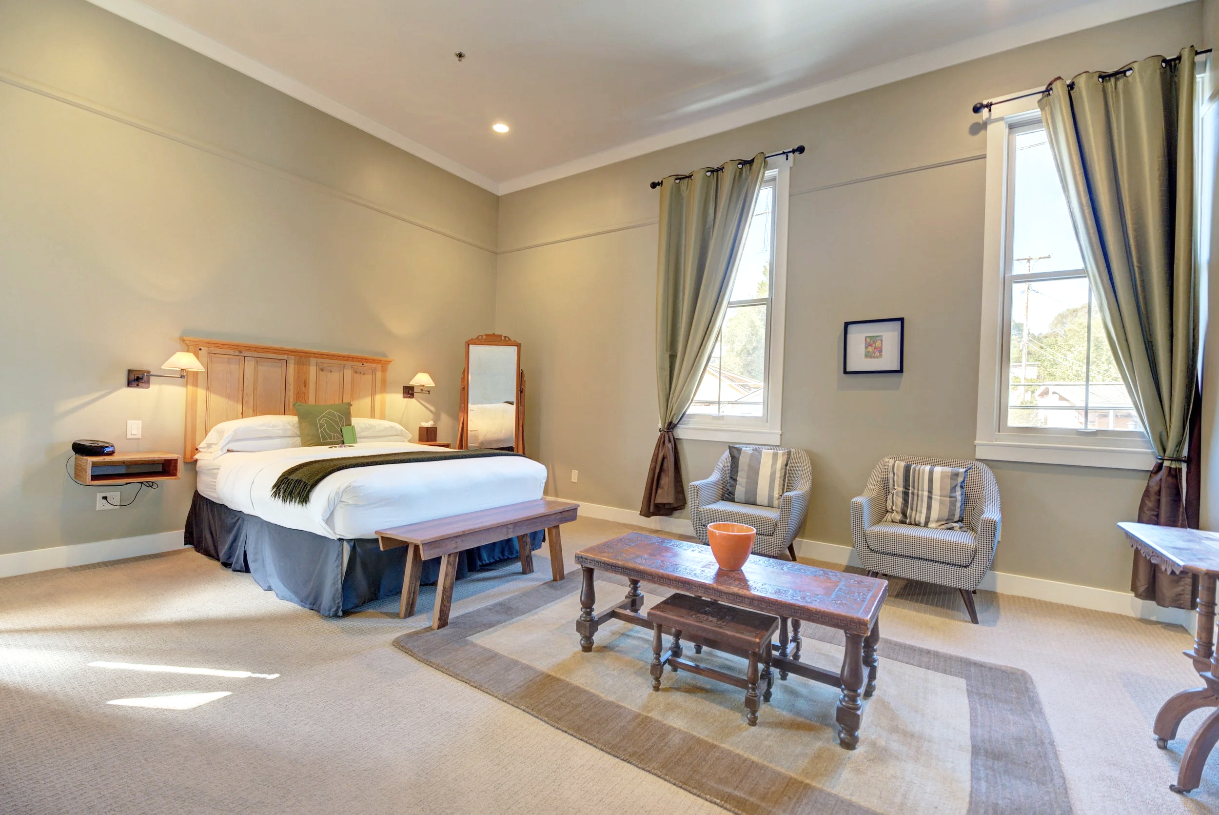 A bedroom with a bed, wooden headboard, two armchairs, a coffee table, and windows with curtains.