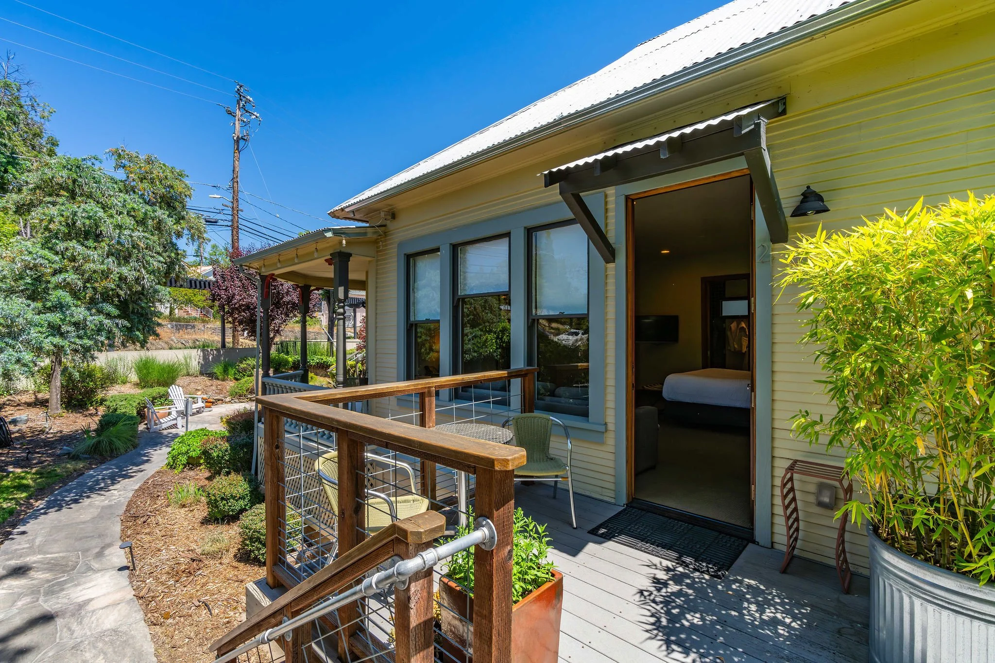 View of a cozy outdoor patio with a wooden railing, surrounded by lush green plants and trees. A pathway leads to a bench in the garden, with a house featuring large windows and an open door outside.