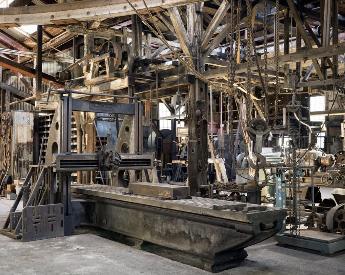 A large vintage industrial machine inside a rusty workshop with metal scaffolding, chains, and pulleys surrounding it.