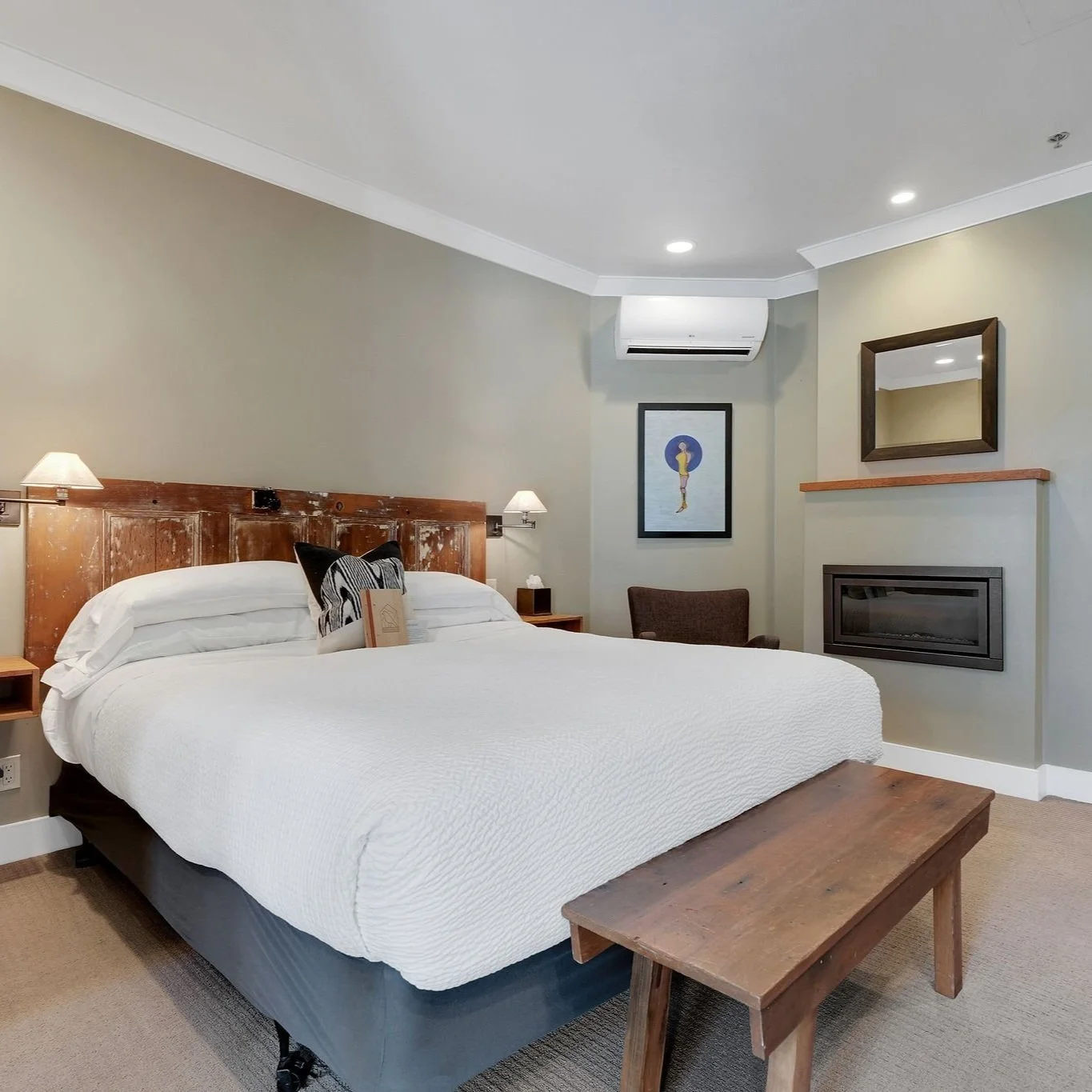 A hotel room with a white bed, wooden headboard, wall-mounted lamps, a small wooden bench at the foot of the bed, a framed artwork on the wall, a chair, a mirror above a fireplace, and an air conditioning unit.