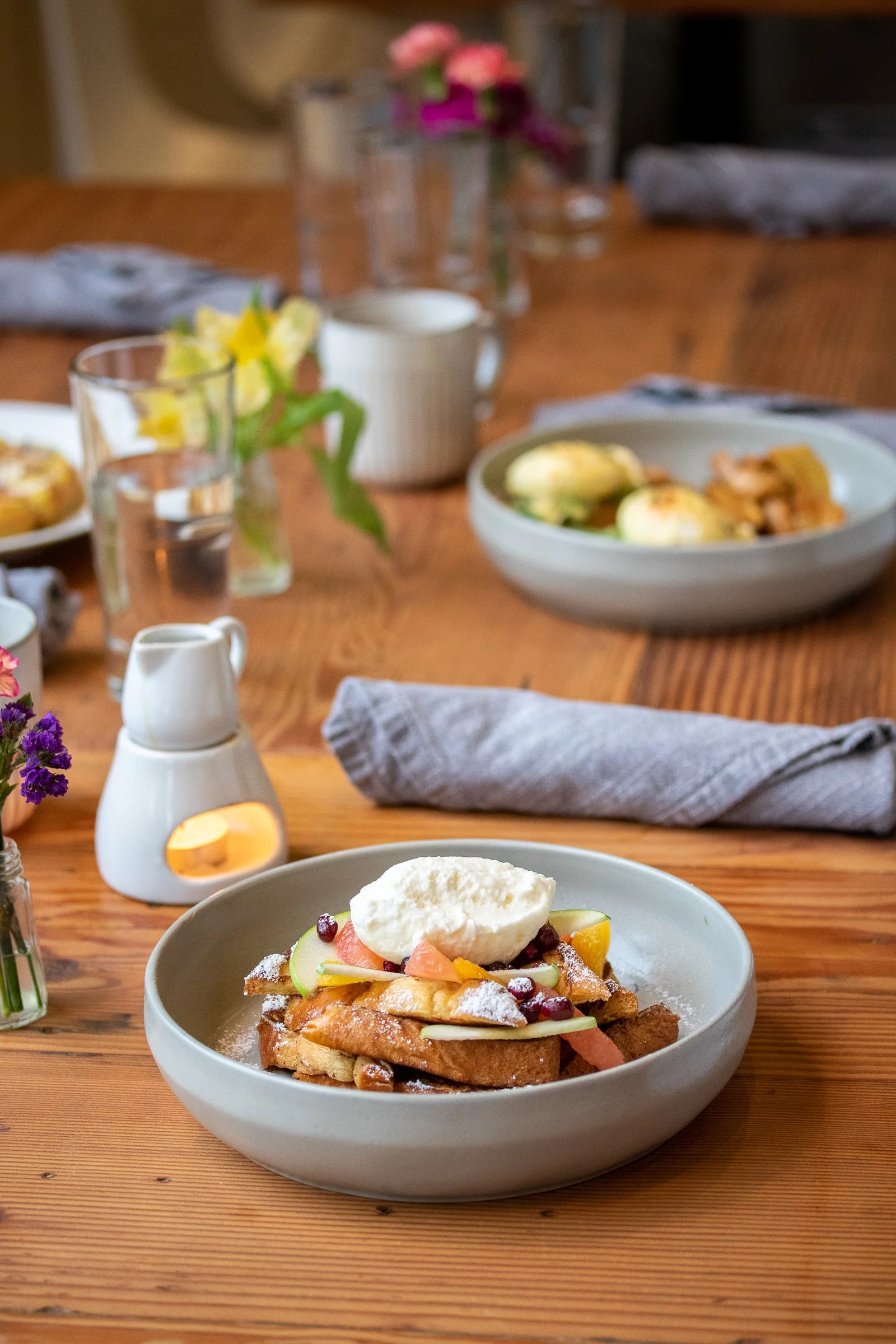 Plate of French toast topped with whipped cream, fruit, and powdered sugar on a wooden table with flowers and drinks