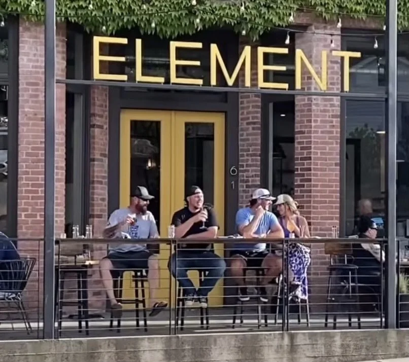 Four people sitting at an outdoor table on a restaurant patio, with a brick building and yellow signage that reads "ELEMENT" behind them.