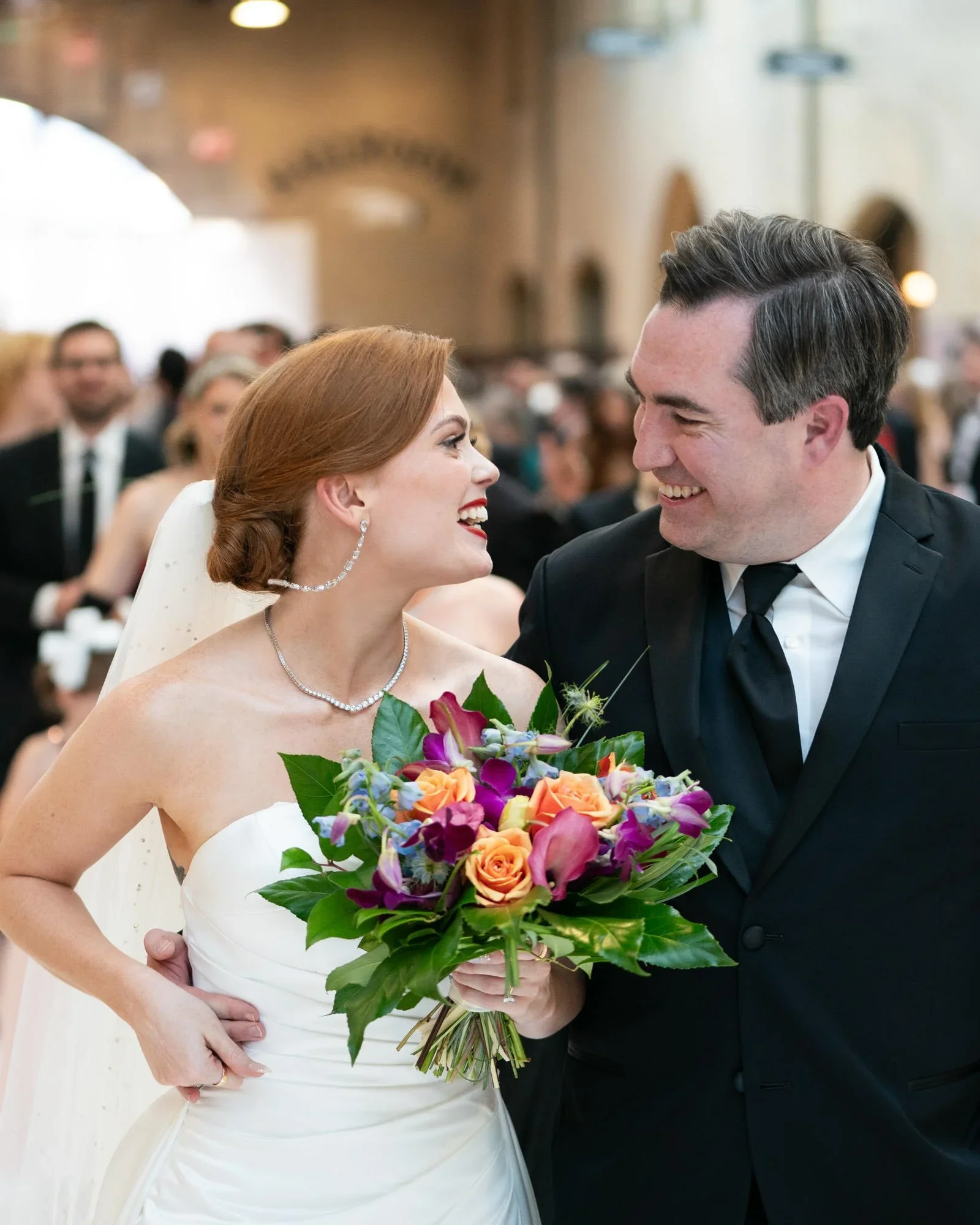 A bride and groom smiling at each other at their wedding, with the bride holding a colorful bouquet of flowers. The bride has red hair and is wearing a white wedding dress with jewelry, while the groom has dark hair and is in a black tuxedo with a white shirt and black tie.