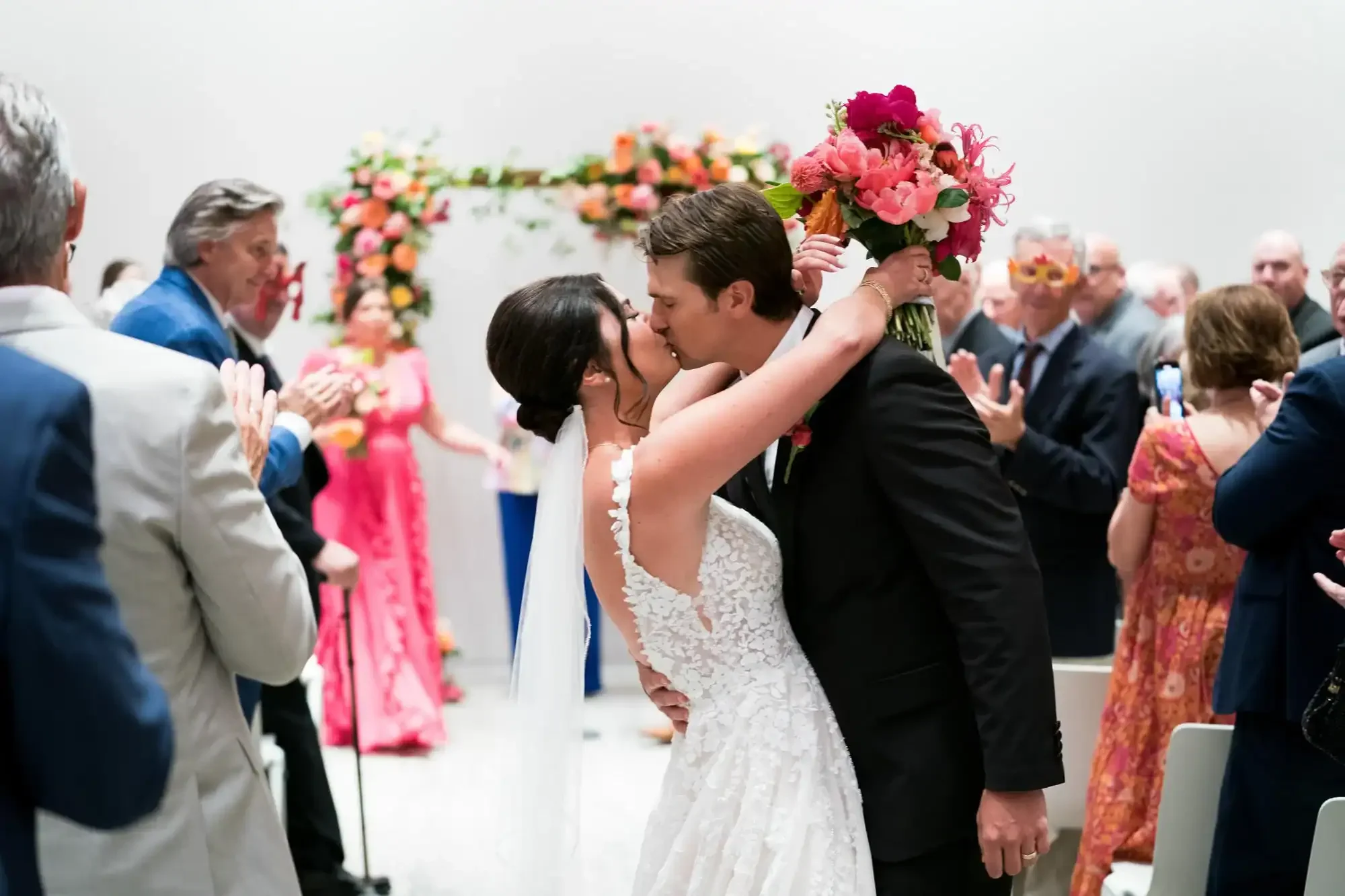 Bride and groom kissing during their wedding ceremony surrounded by guests, with the bride holding a bouquet of pink and red flowers, and an arch decorated with flowers in the background.
