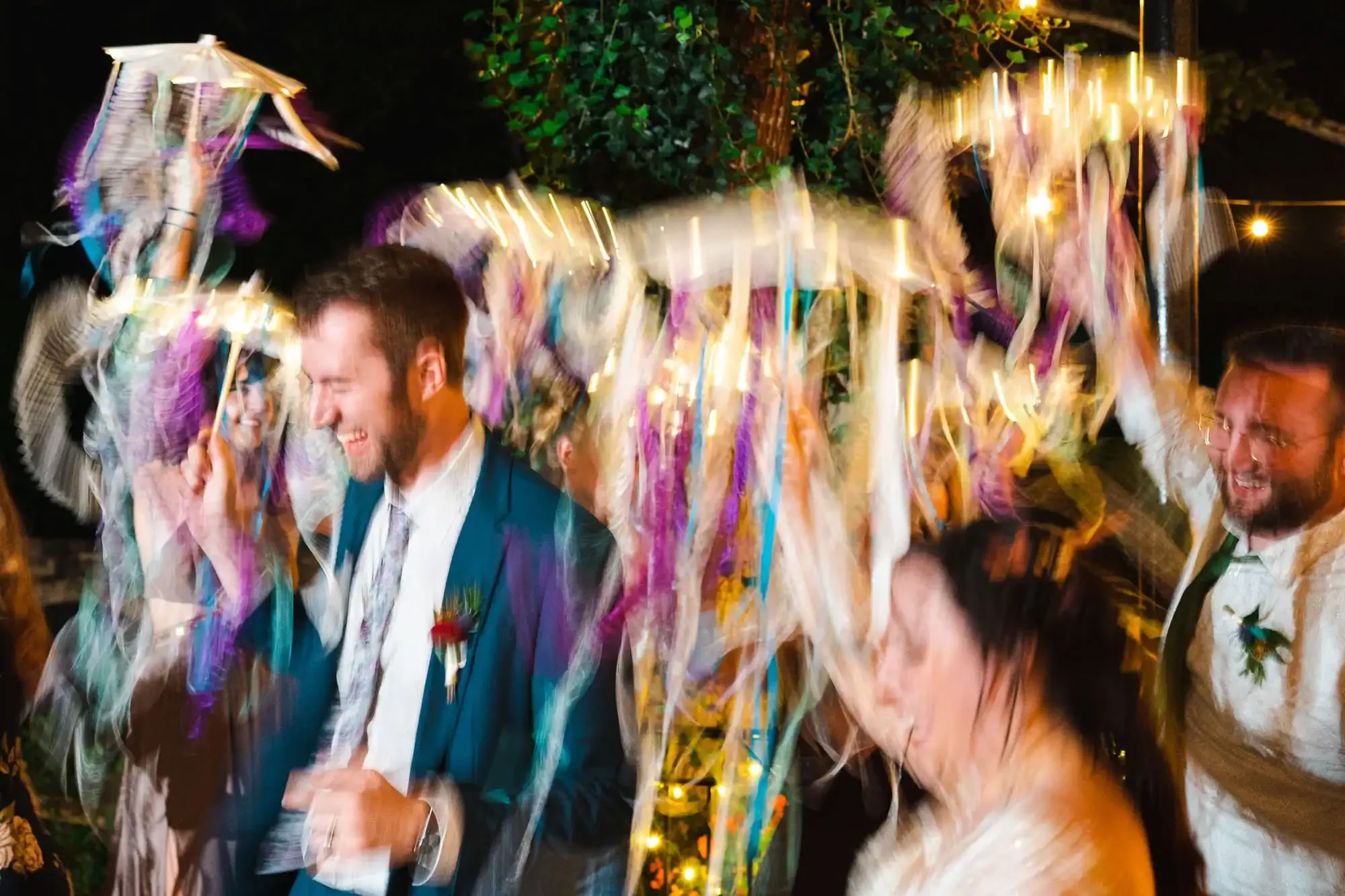 A group of people at a celebration or wedding, holding umbrellas and smiling, with colorful streamers and lights around them.