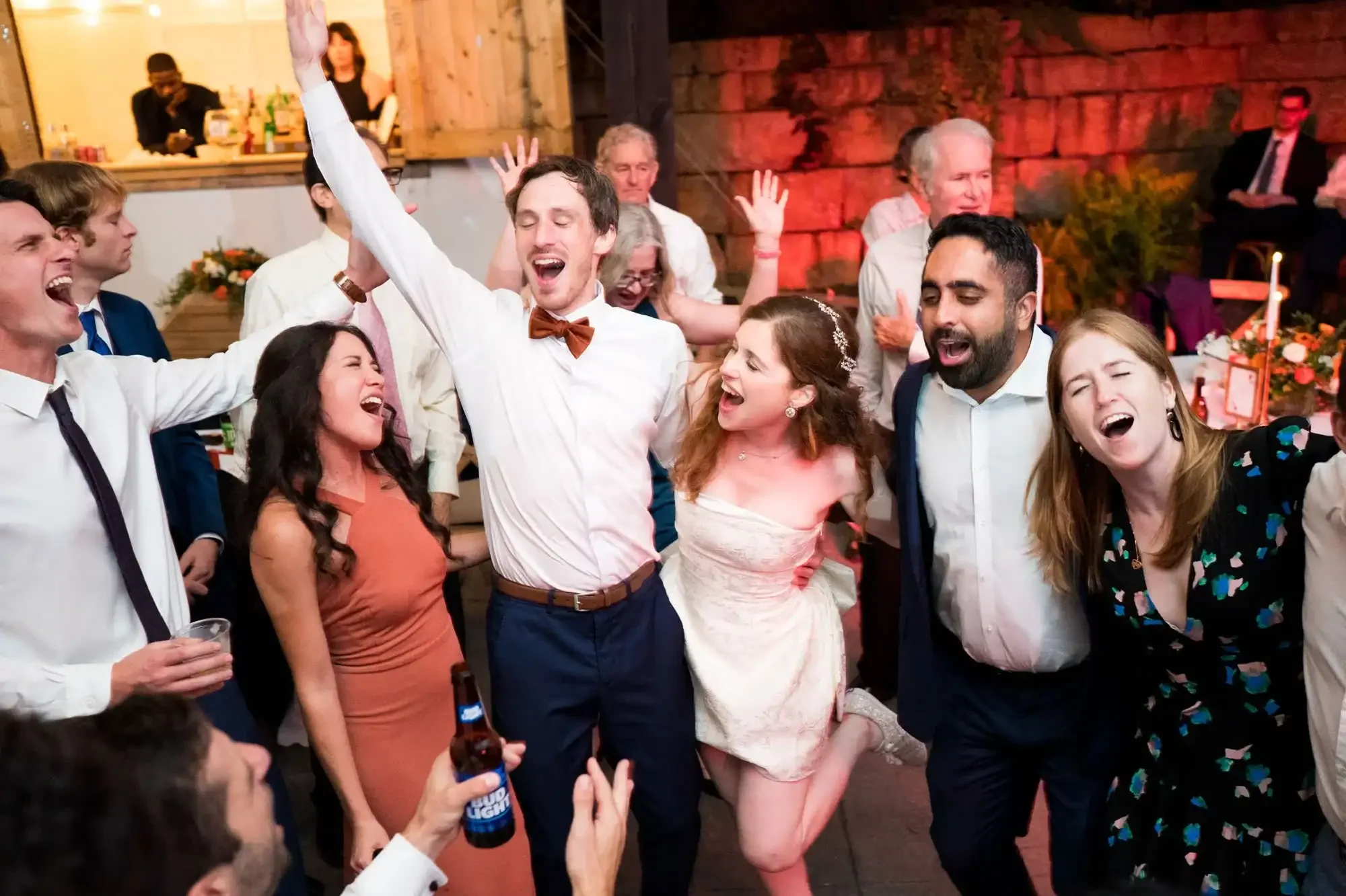 A group of people celebrating and dancing at a wedding reception, with some raising their hands and singing.
