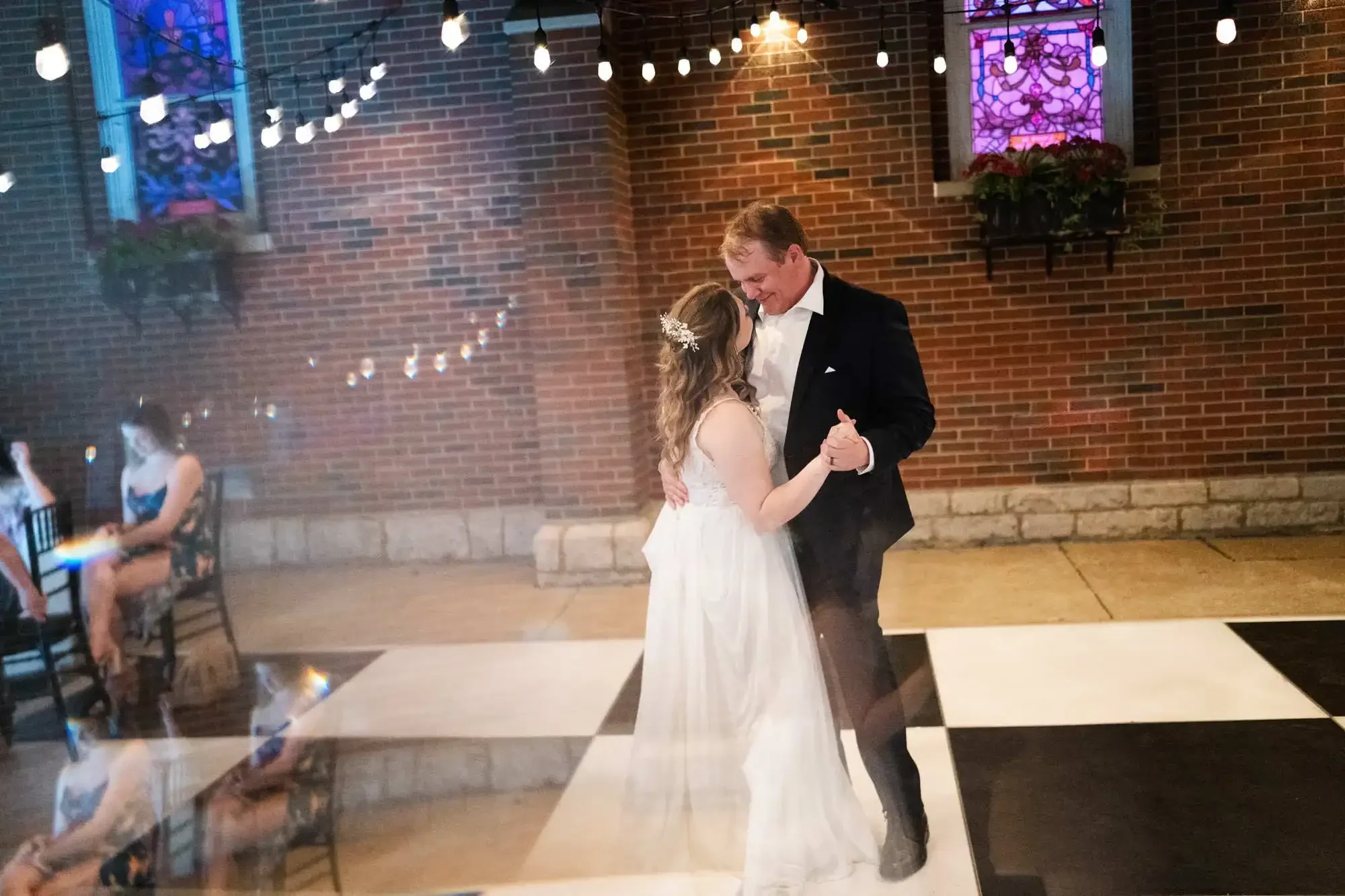 A bride and groom sharing their first dance at their wedding reception, with string lights overhead, a brick wall background, and stained glass windows.