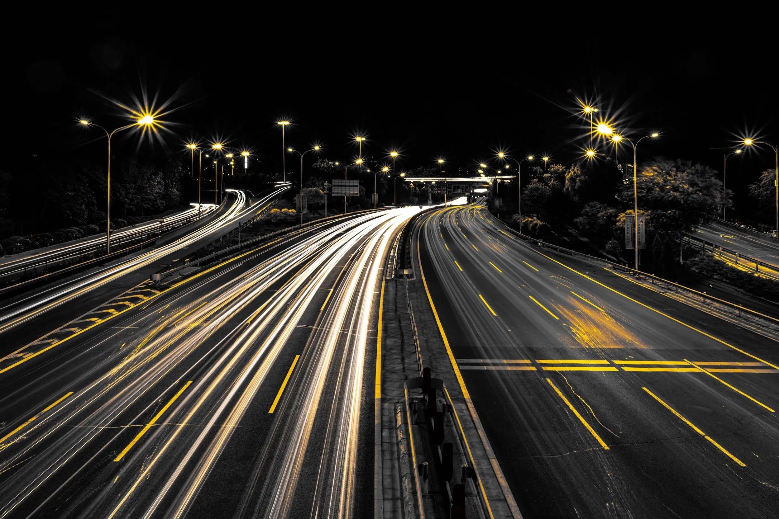 Nighttime long exposure photograph of a highway with streaks of light from moving vehicles, illuminated streetlights, and curving lanes.