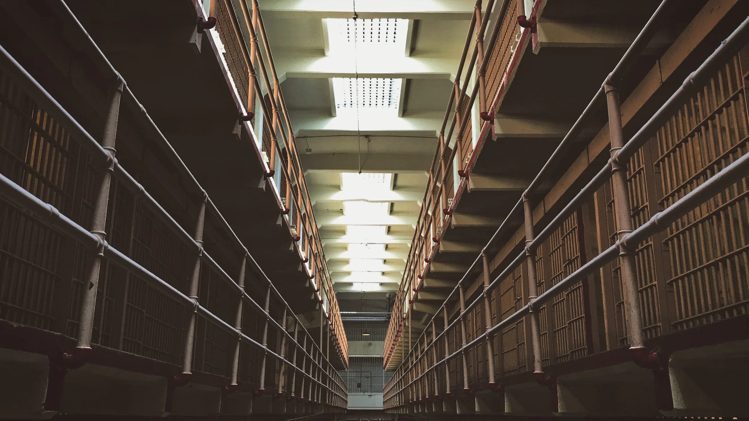 Interior view of a prison with two levels of cell blocks, metal railings, and fluorescent ceiling lights.