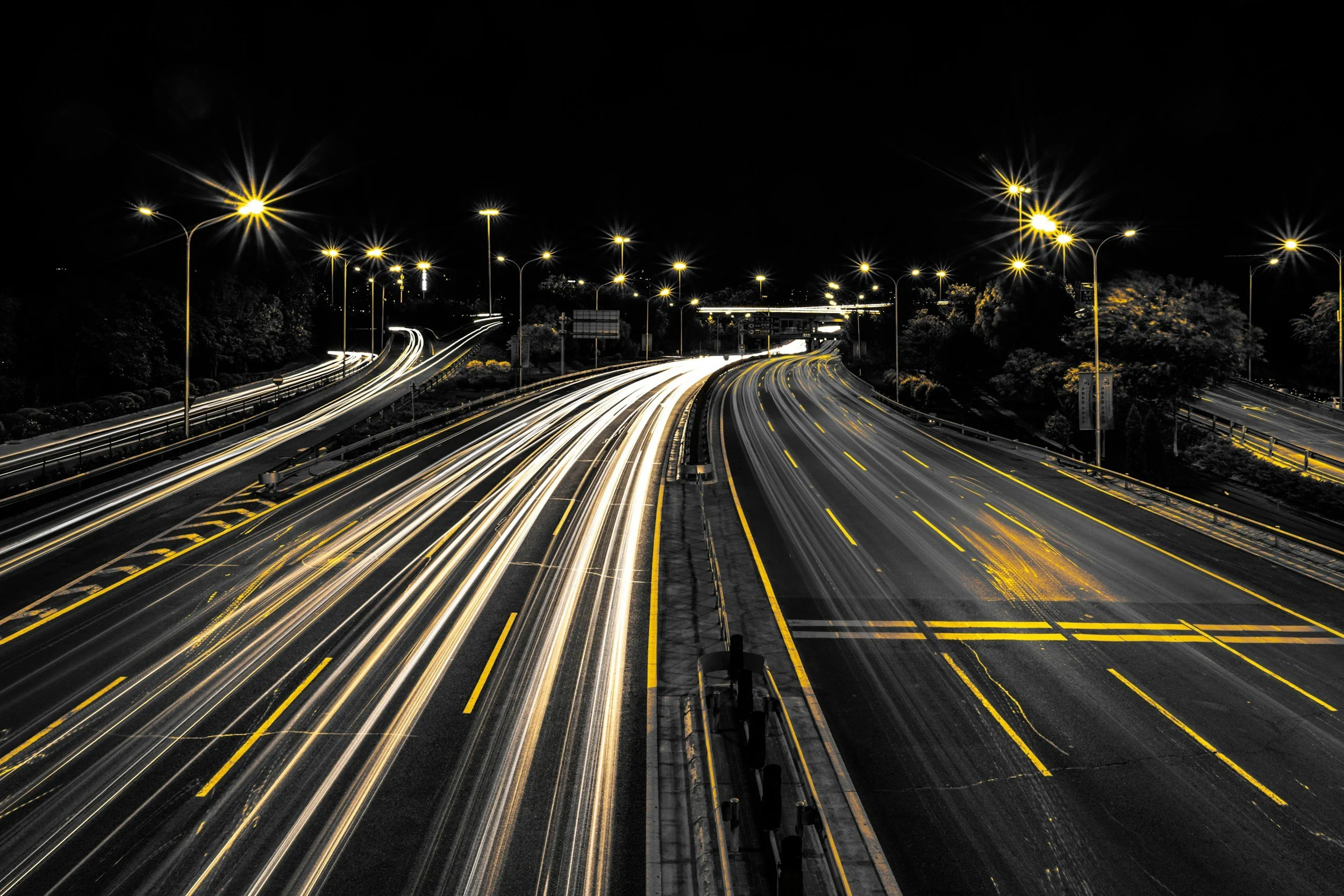 Long exposure photo of a multi-lane highway at night with streaks of car headlights and taillights, illuminated streetlights, and trees on the sides.