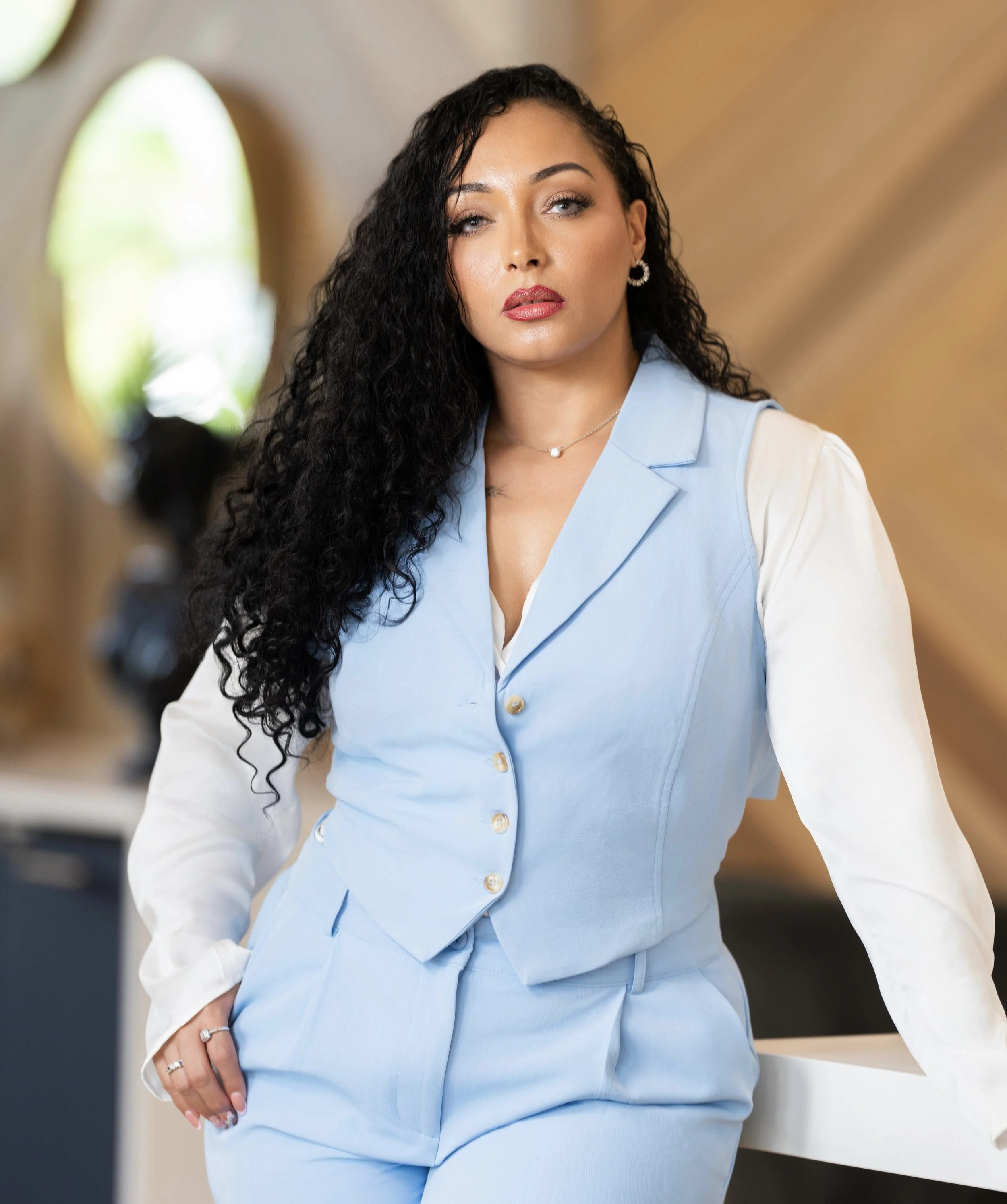 A woman with long curly black hair, wearing a light blue suit with a white shirt, standing indoors with a blurred background and arched window.