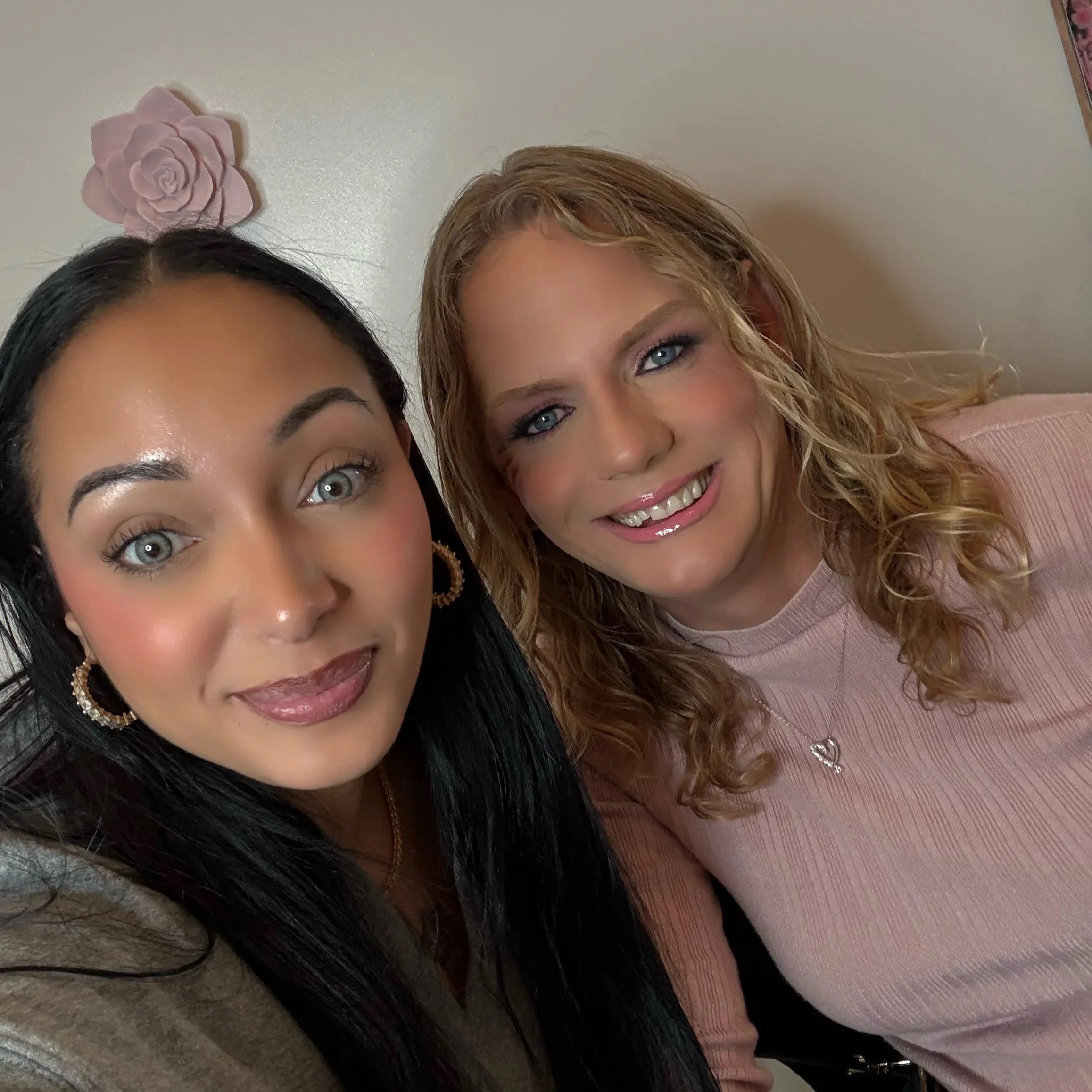 Two women smiling close together, one with dark hair and the other with curly blonde hair, with a pink wall and pink flower decoration behind them.