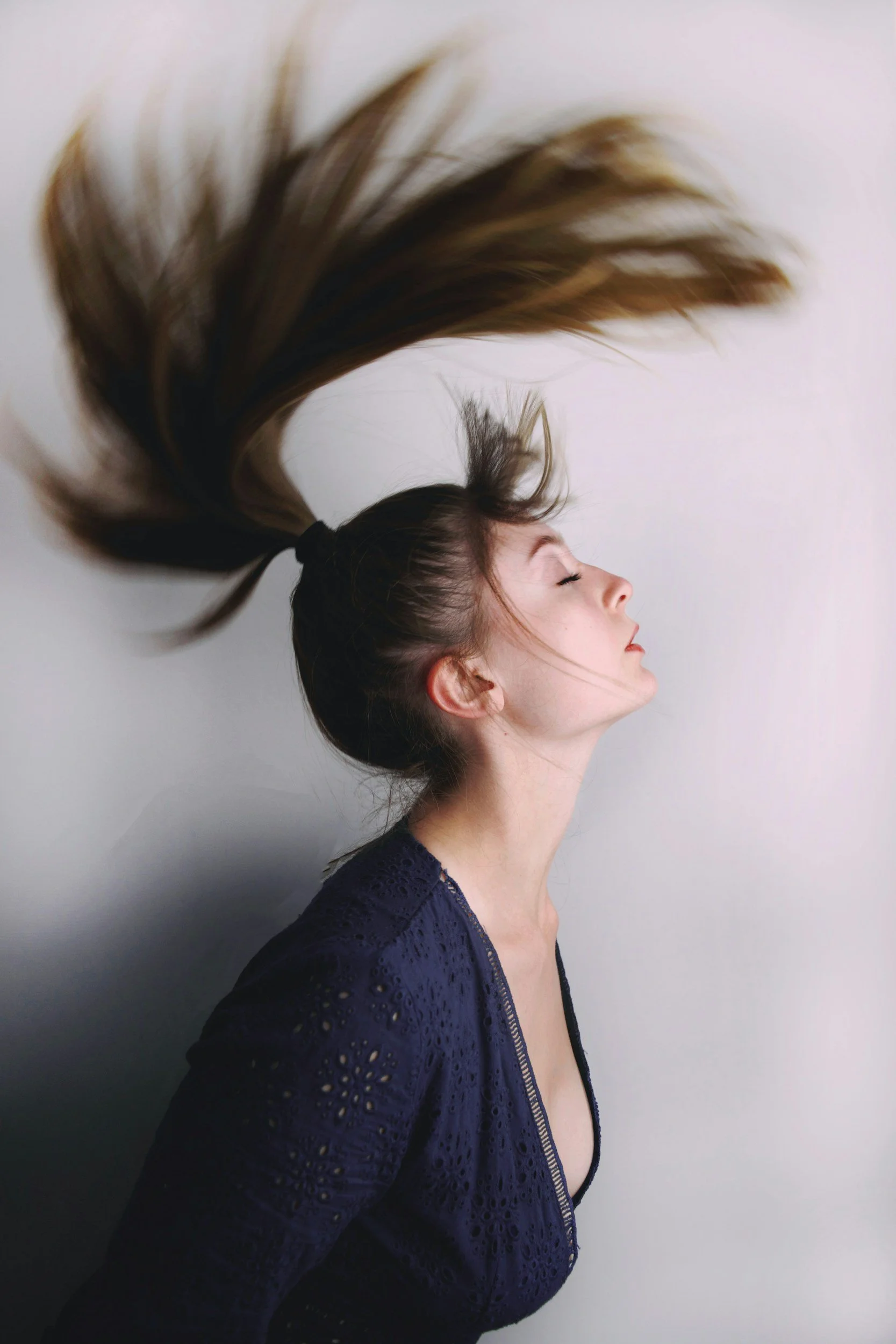 A woman with long brown hair in a ponytail flipping her hair, wearing a dark blue lace top, with closed eyes and a light background.