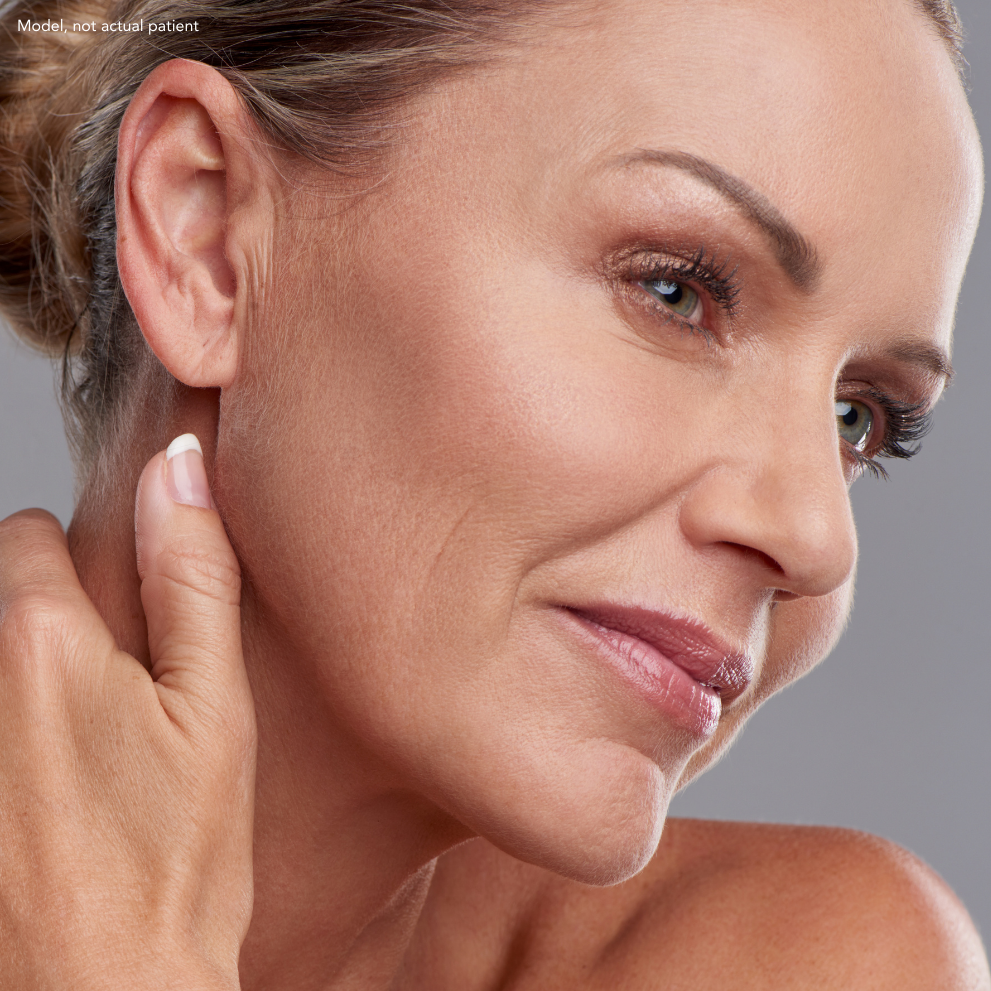 Close-up of a middle-aged woman touching her neck, with smooth skin, makeup, and gray background.