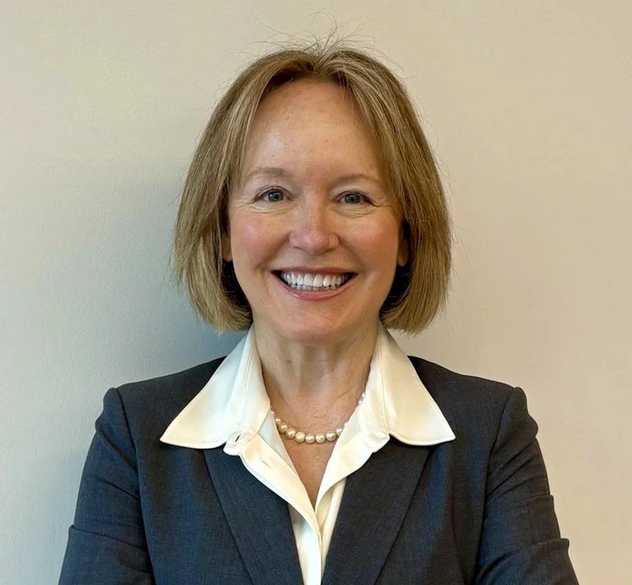 Headshot of a smiling woman with blonde, wavy hair, wearing a black blazer and white shirt, against a gray background.
