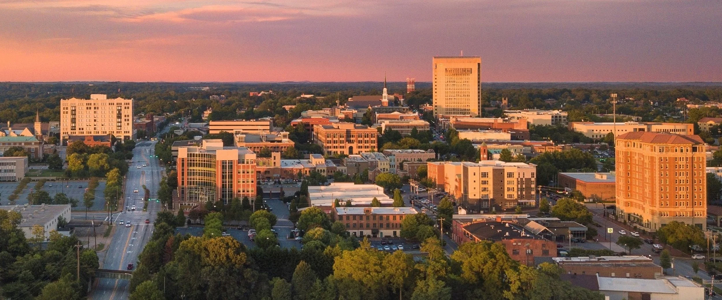 Aerial view of a cityscape at sunset with tall buildings, trees, and roads.