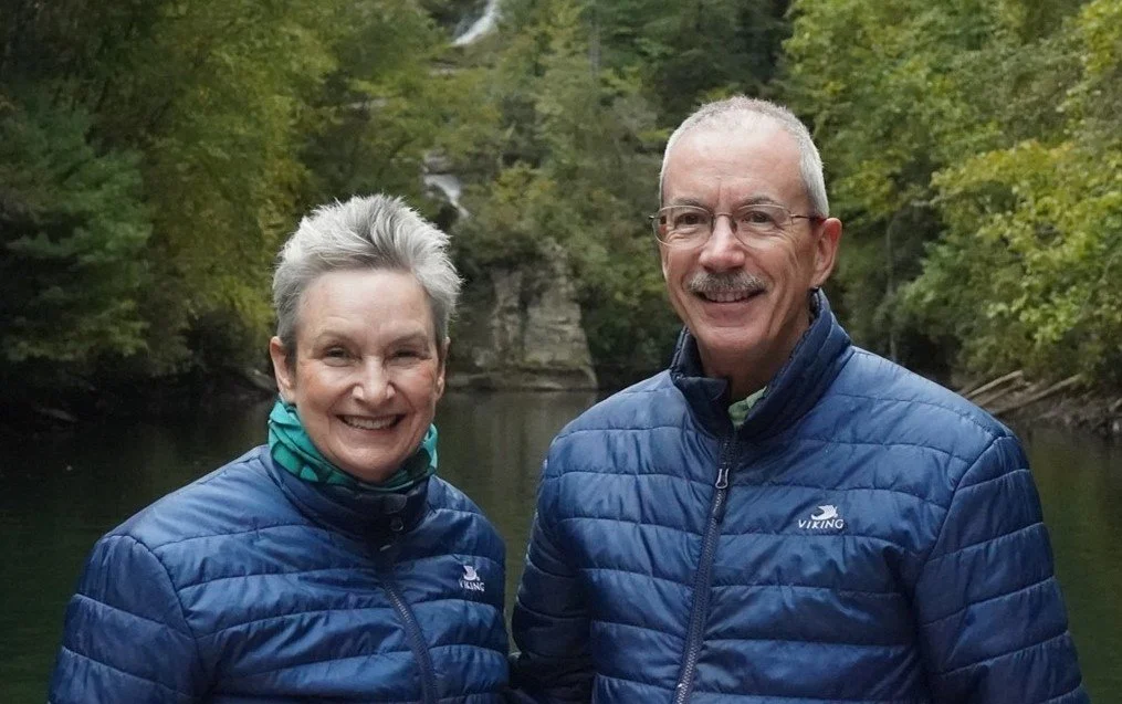 A smiling elderly couple outdoors on a river with trees in the background.