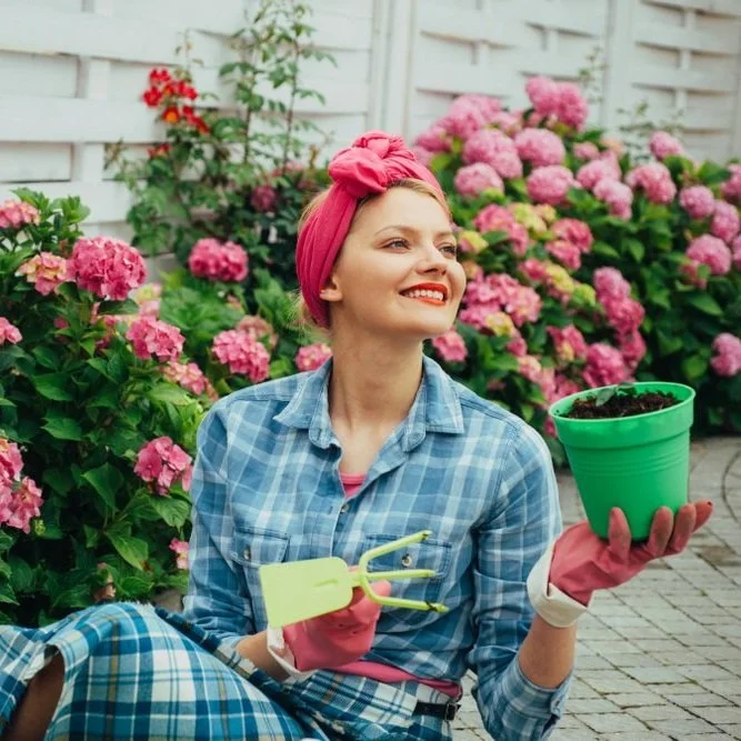 Woman gardening holding green pot and yellow shovel. Pink flowers in the background.