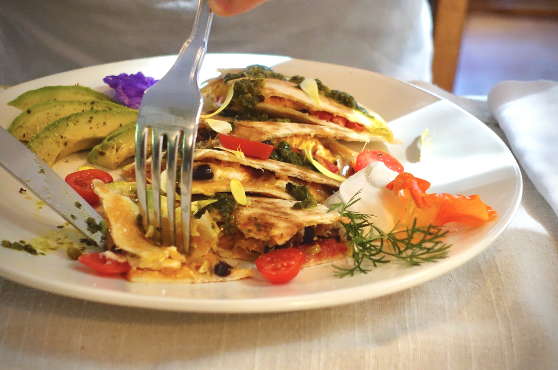 A white plate with quesadilla topped with green sauce, cherry tomatoes, and garnished with herbs. Sliced avocado with black pepper, purple edible flowers and a dollop of sour cream with orange edible flower petals sprinkled on top.