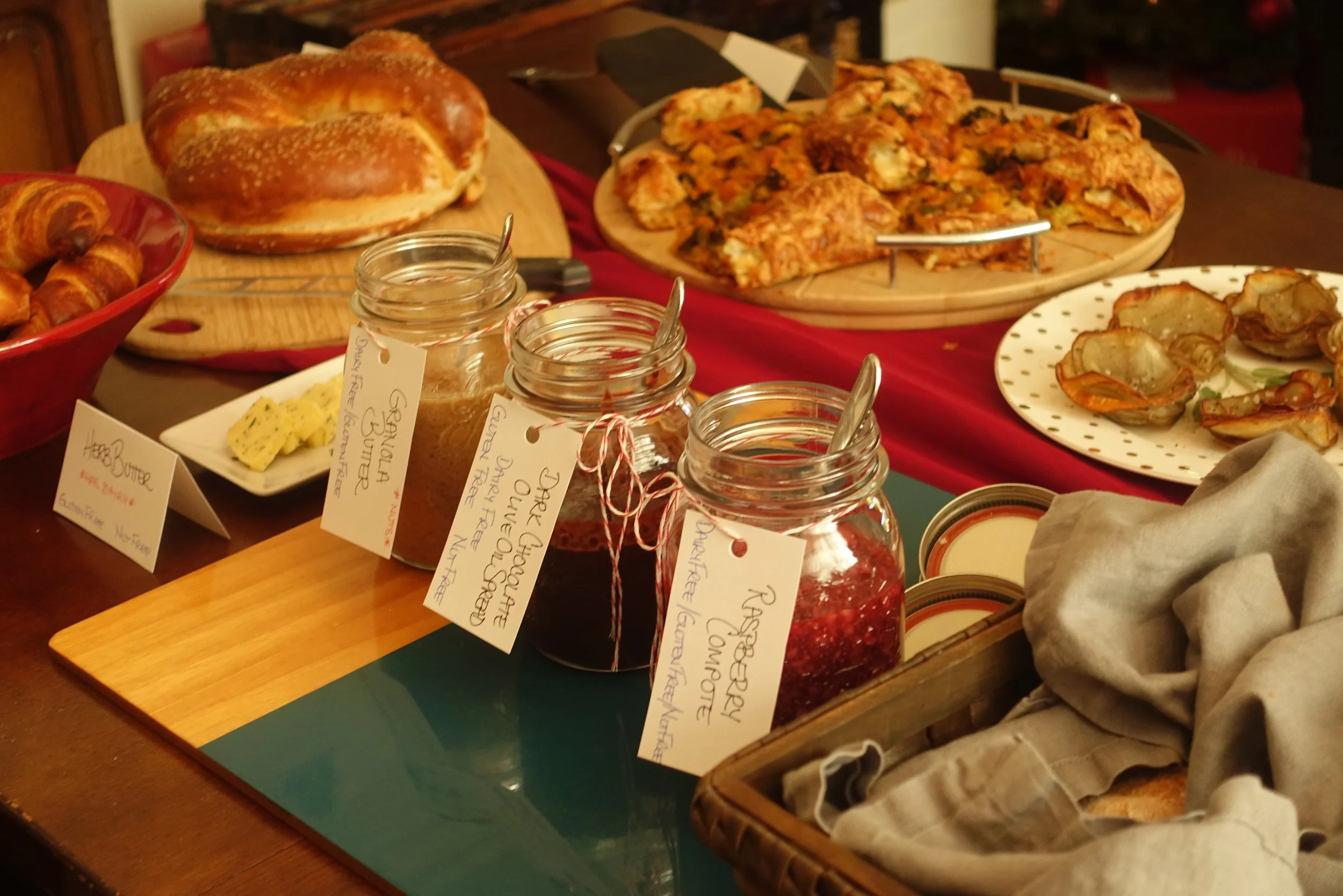A spread of breakfast foods including croissants, a loaf of bread, a pizza, baked eggs, and jars of jam and butter on a table with handwritten labels