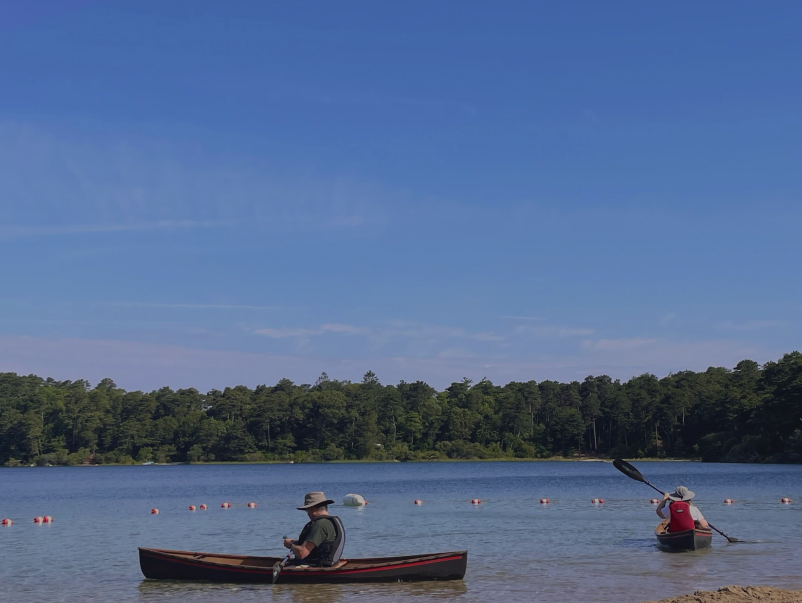 Two people in canoes on a body of water, with a tree-lined shoreline in the background and a clear blue sky overhead.