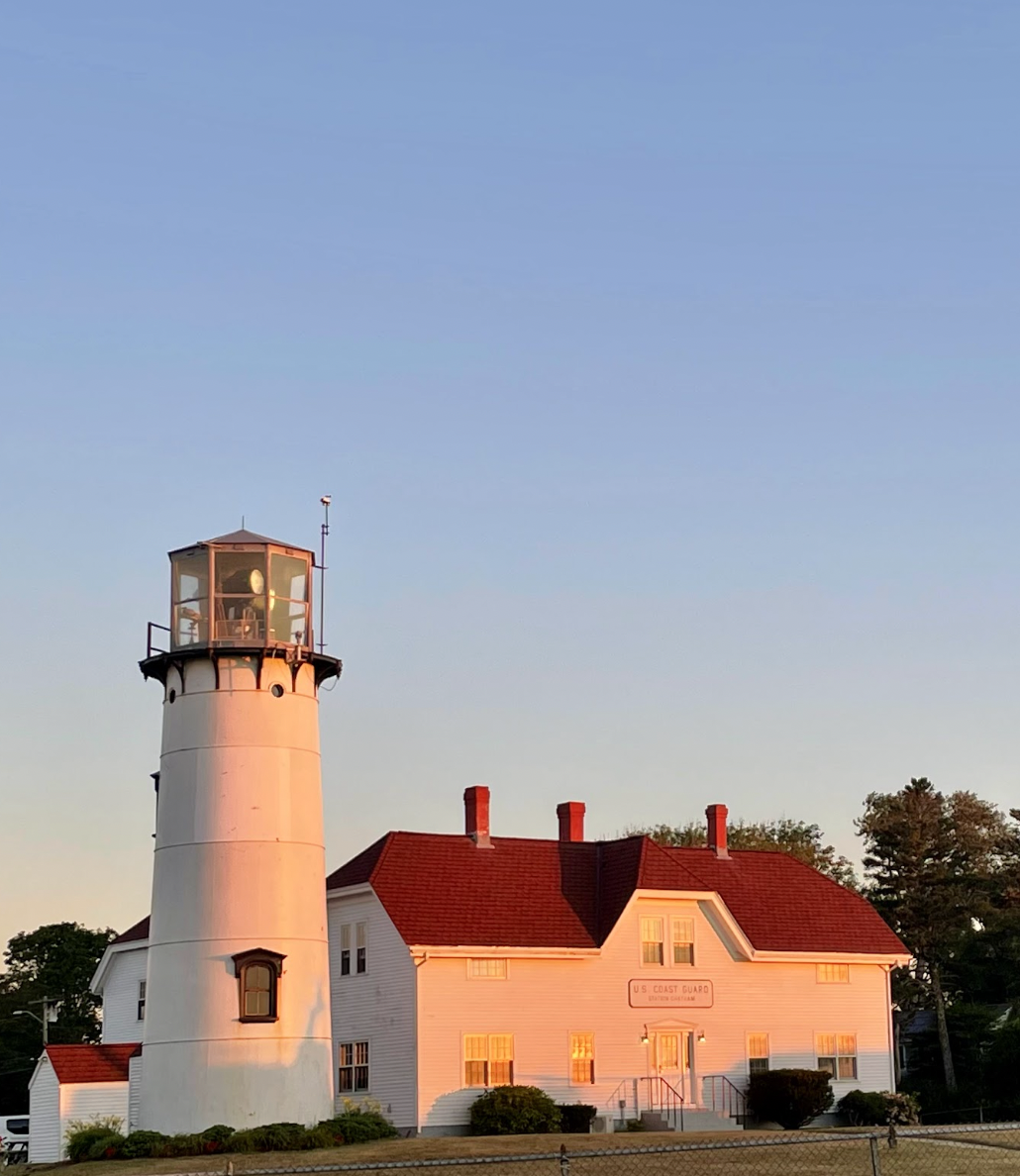 A white lighthouse with a red roof adjacent to a white building labeled 'U.S. Coast Guard' with several windows, set against a clear blue sky during sunset.