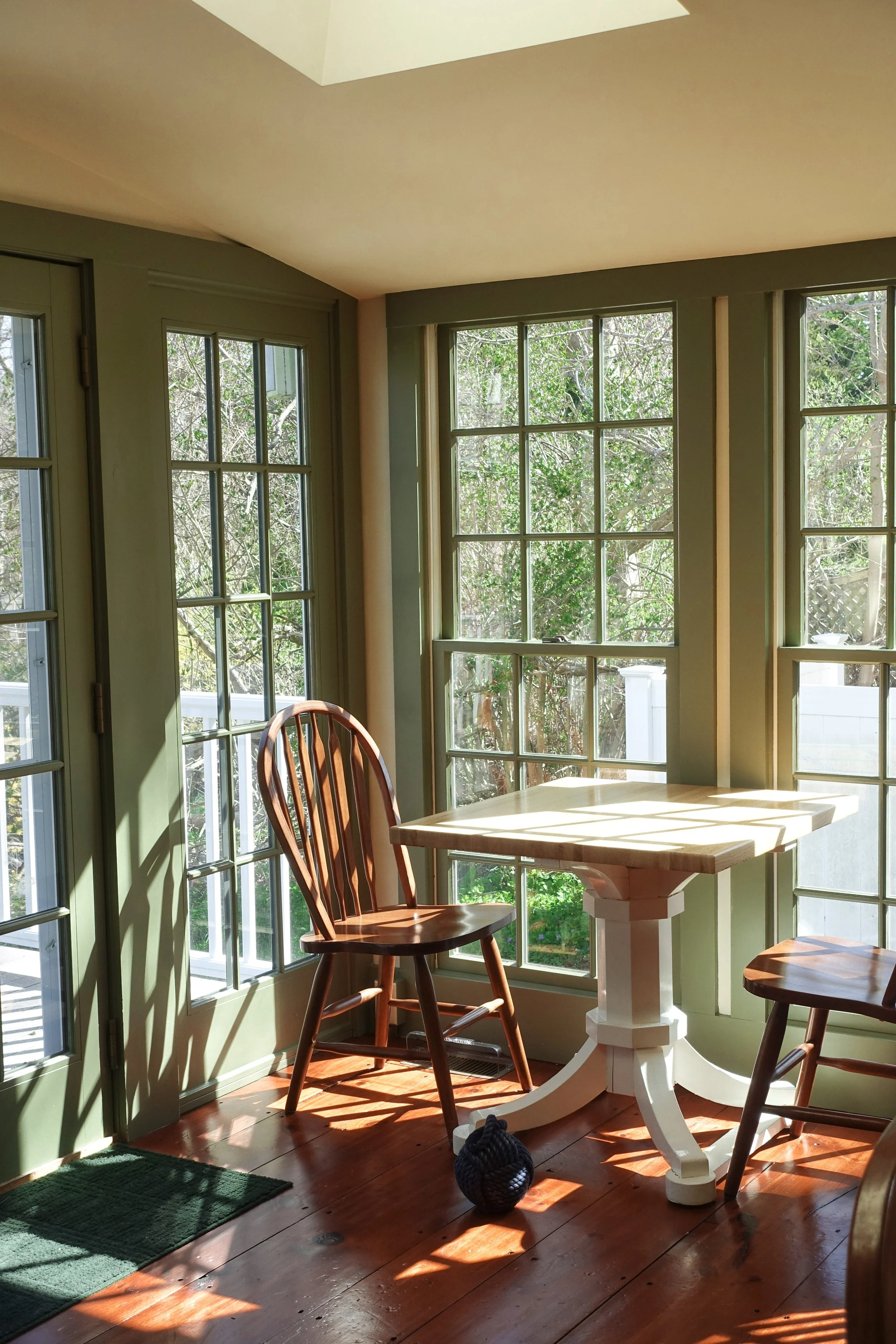 Sunlight streams through large window panes illuminating a cozy corner with a wooden table, two wooden chairs, a small decorative vase, and a green rug on a wooden floor.