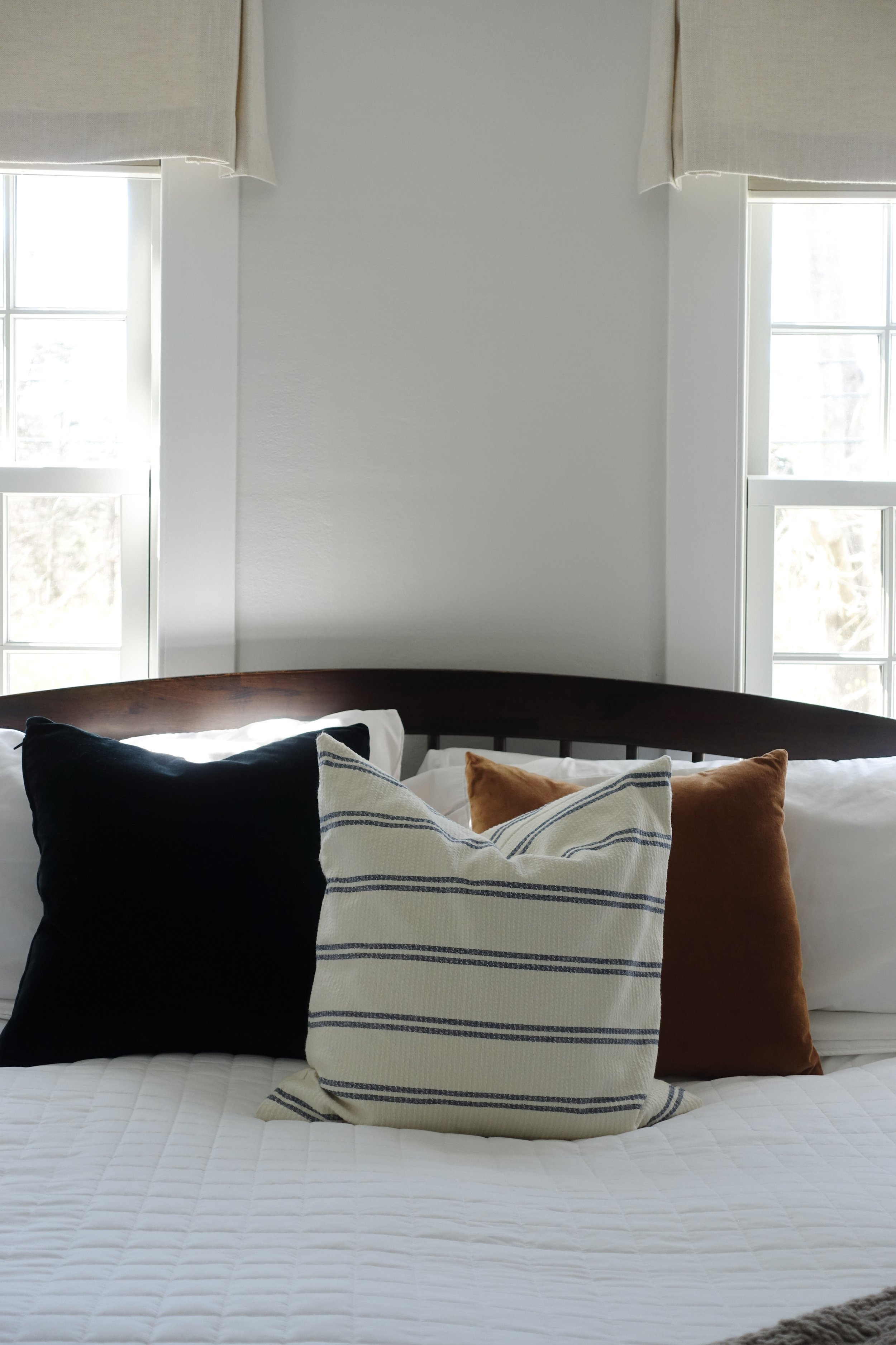 A neatly made bed with a white quilt, three pillows (black, striped beige and blue, and brown) against a headboard, in front of two windows with beige curtains, in a bright room.